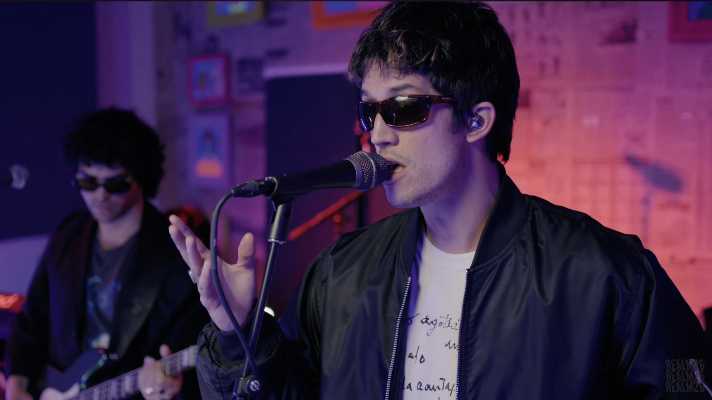 A singer wearing sunglasses and a black jacket singing into a microphone, with a guitarist in the background in a brightly colored room.