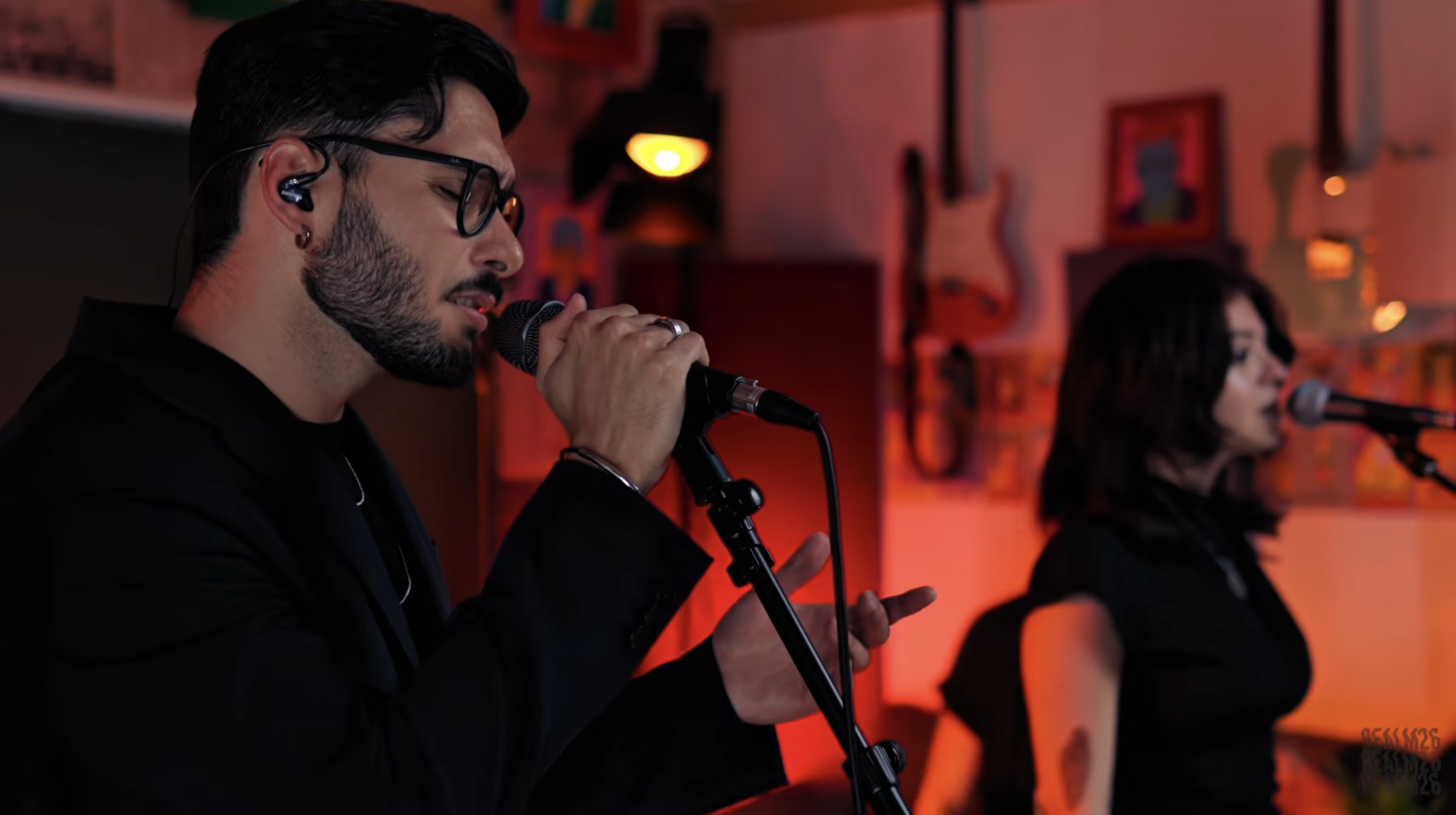A man and woman with microphones singing in a dimly lit music studio with guitars and colorful art on the walls.