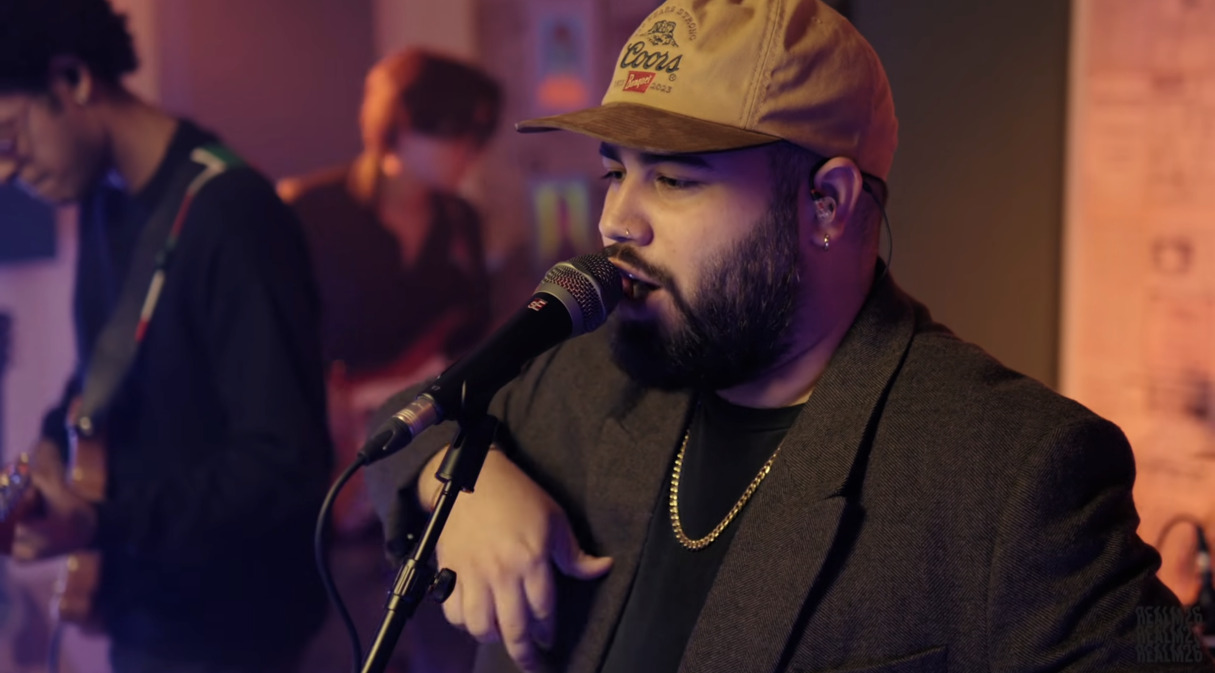 A man with a beard, wearing a tan cap and gold chain, sings into a microphone in a dimly lit bar or club, with other musicians in the background.