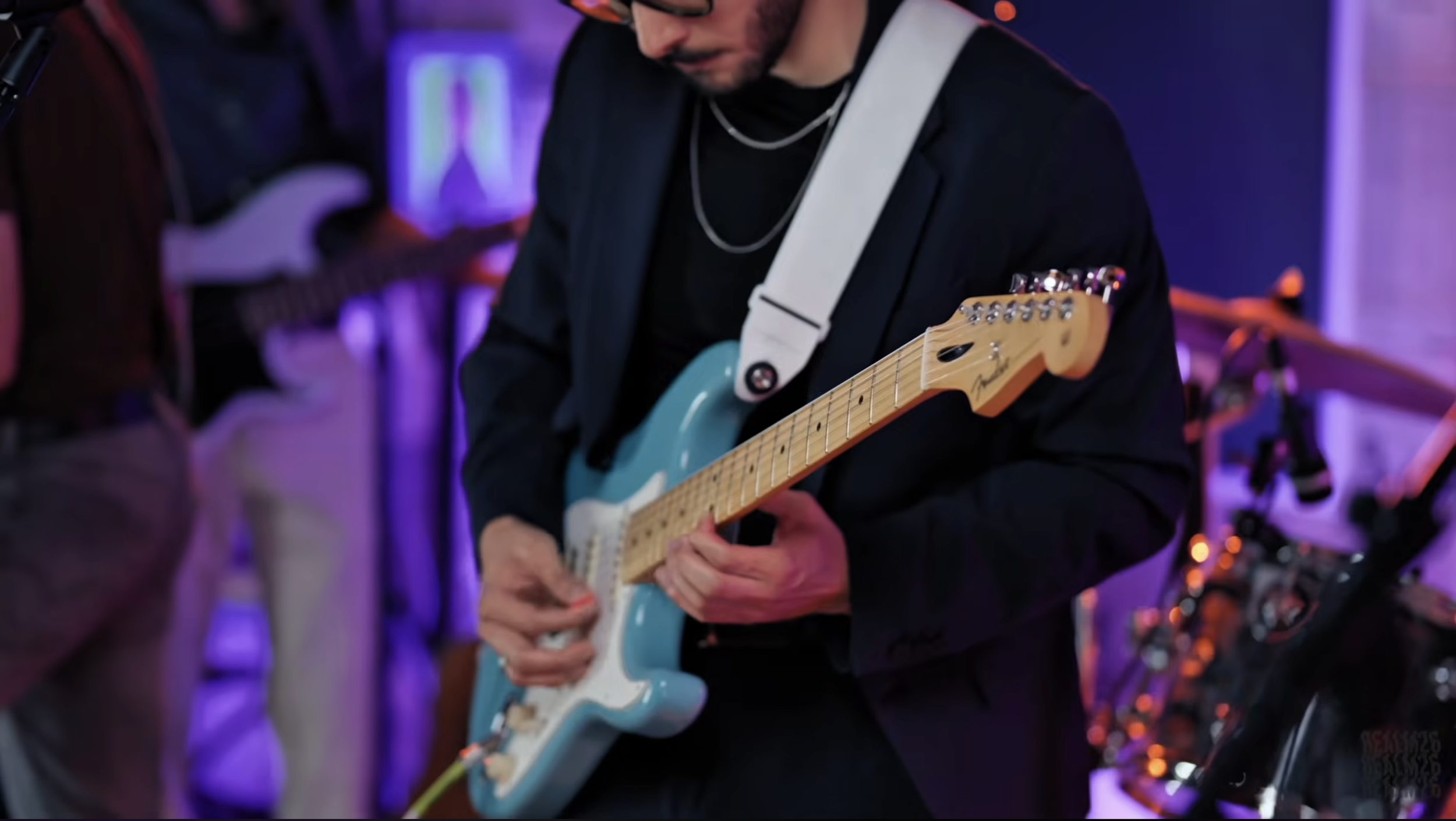 Man playing a blue electric Fender Stratocaster guitar in a dimly lit room with purple and orange lighting.