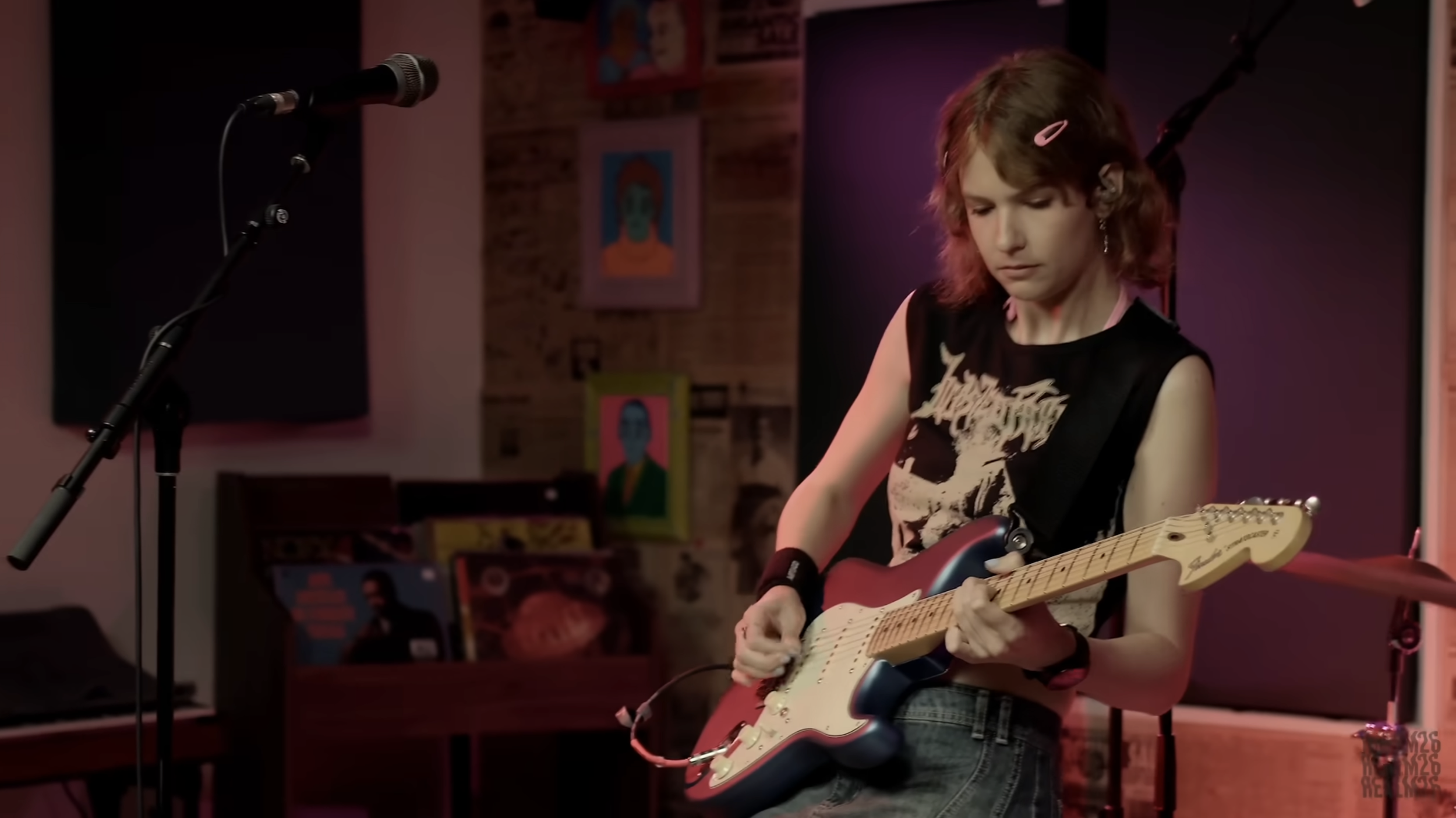 A young woman with wavy brown hair wearing a black sleeveless shirt playing an electric guitar in a dimly lit music studio or performance space.
