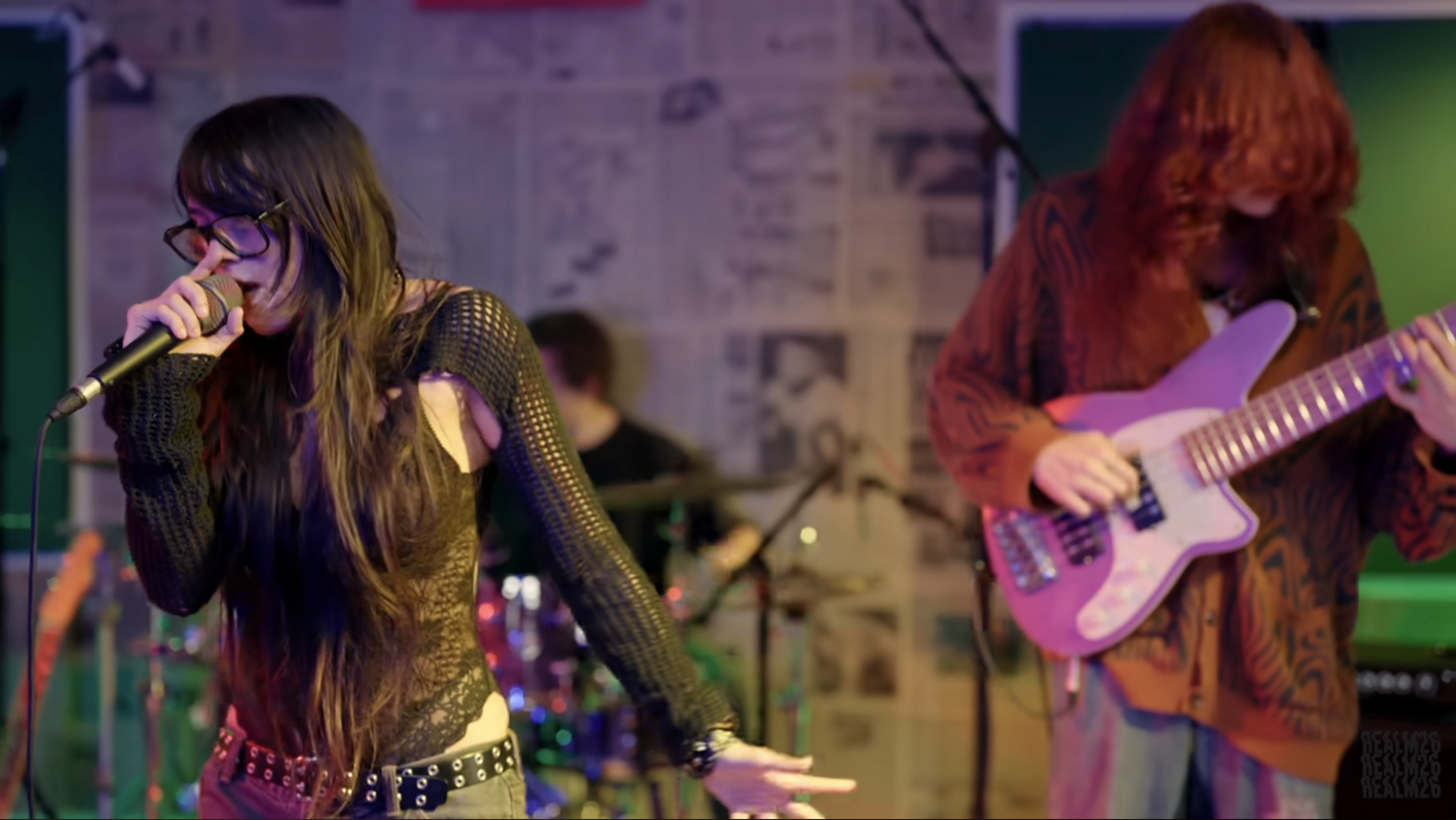 A music band performing in a venue with a female singer in a black lace top and glasses holding a microphone, a male guitarist in a brown jacket playing a purple guitar, and a drummer in the background.