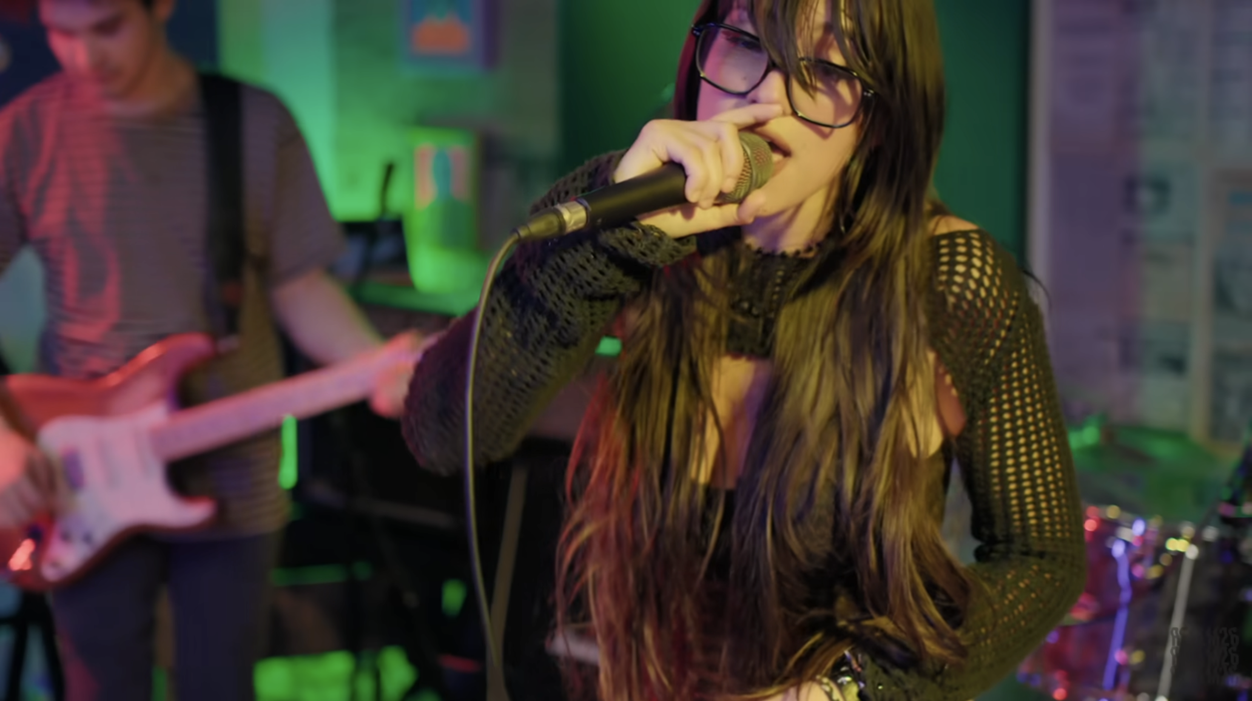 A young woman with long hair and glasses singing into a microphone during a musical performance in a colorful, illuminated setting with a guitarist in the background.