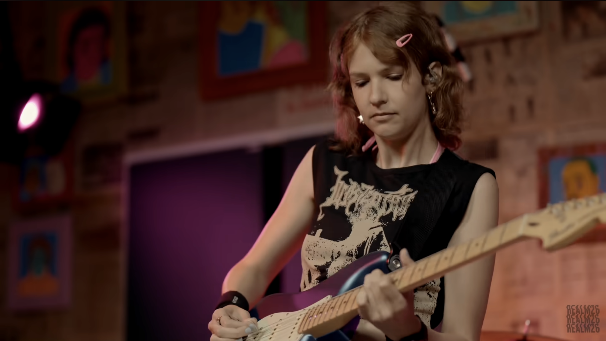A young girl with shoulder-length red hair playing an electric guitar on a stage with a brick wall background and colorful wall art.