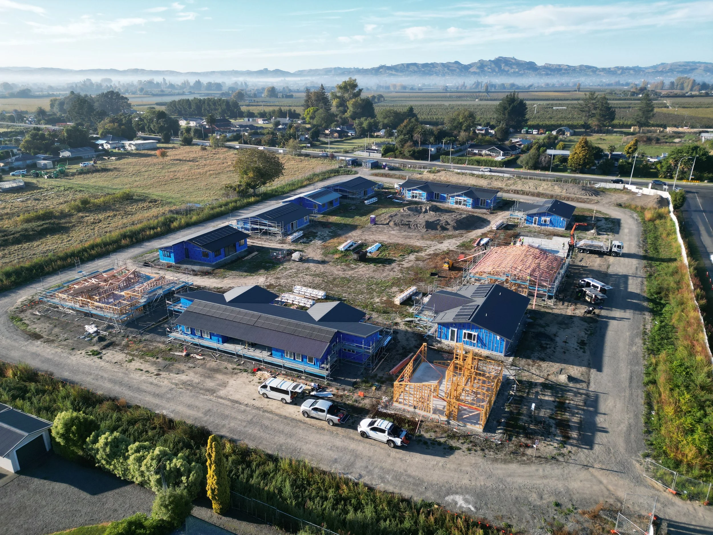 Aerial view of a suburban construction site with multiple houses being built, surrounded by fields and countryside.