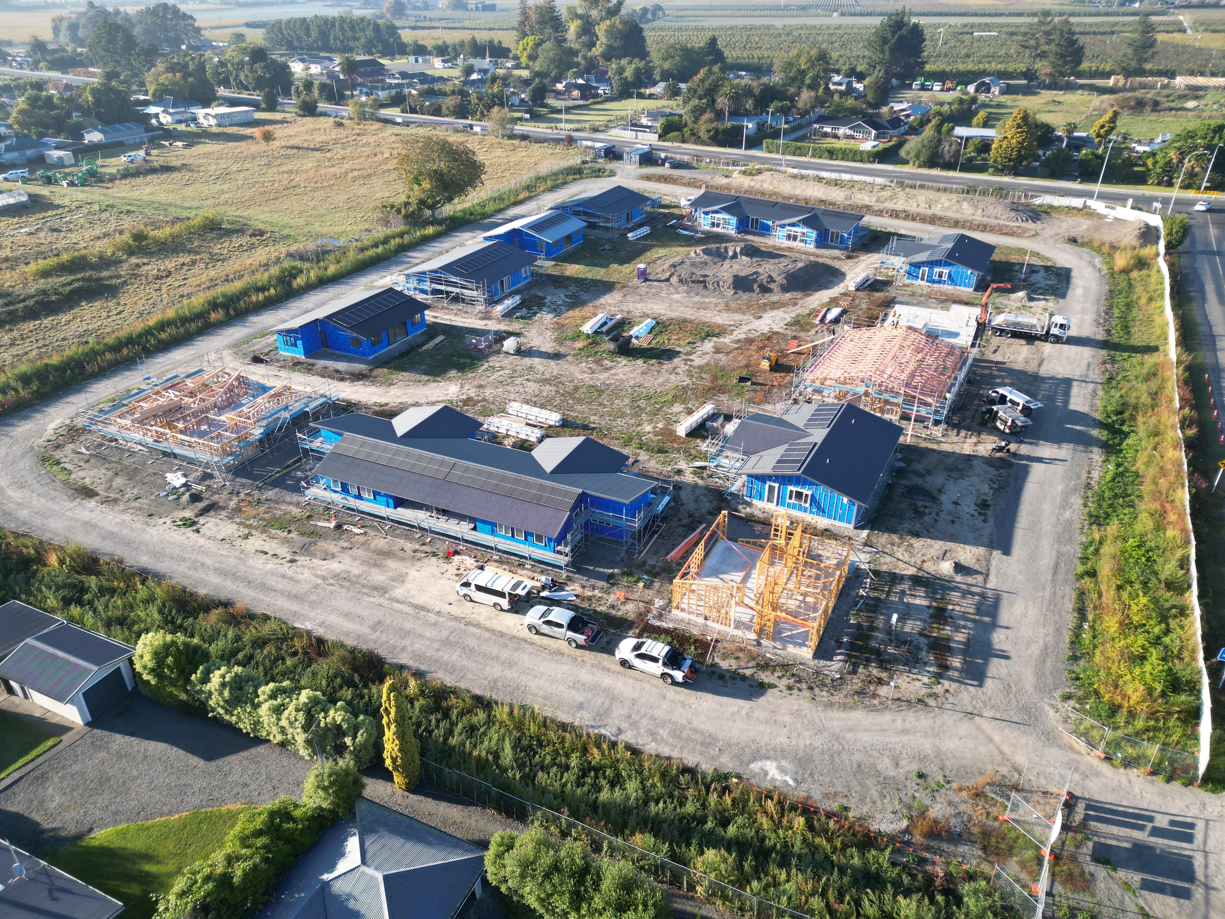 Aerial view of a residential construction site with multiple buildings in various stages of development, surrounded by fields and roads.