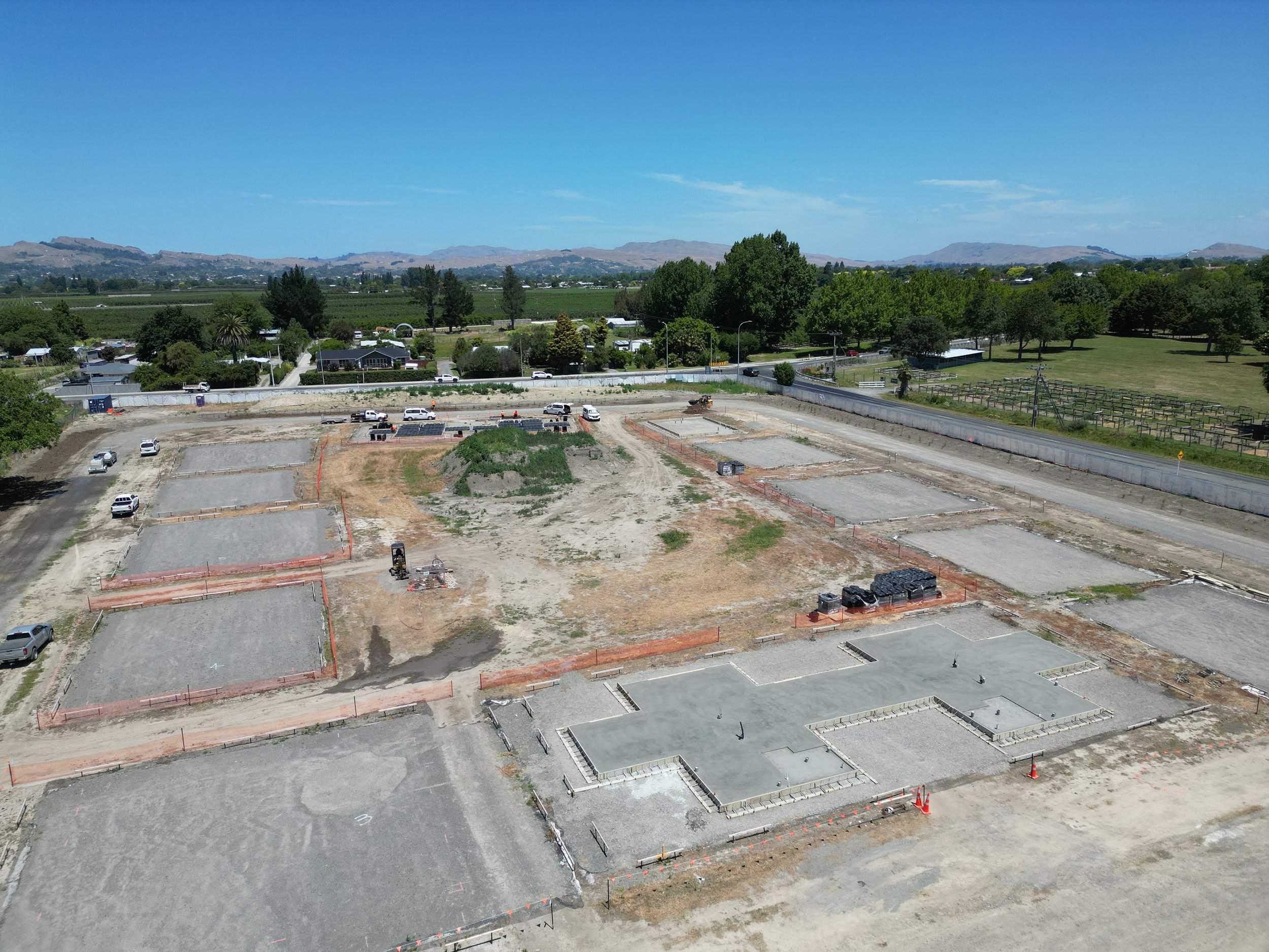 A construction site with multiple outlined building foundations, surrounded by dirt and construction equipment. Several vehicles are parked around the site. In the background, there are trees, a road, and distant hills under a clear blue sky.