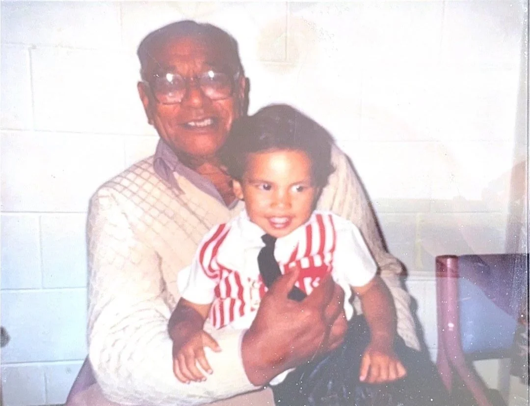 Elderly man smiling and holding a young child, who is wearing a white shirt with red stripes and a black tie. The background features a white brick wall.