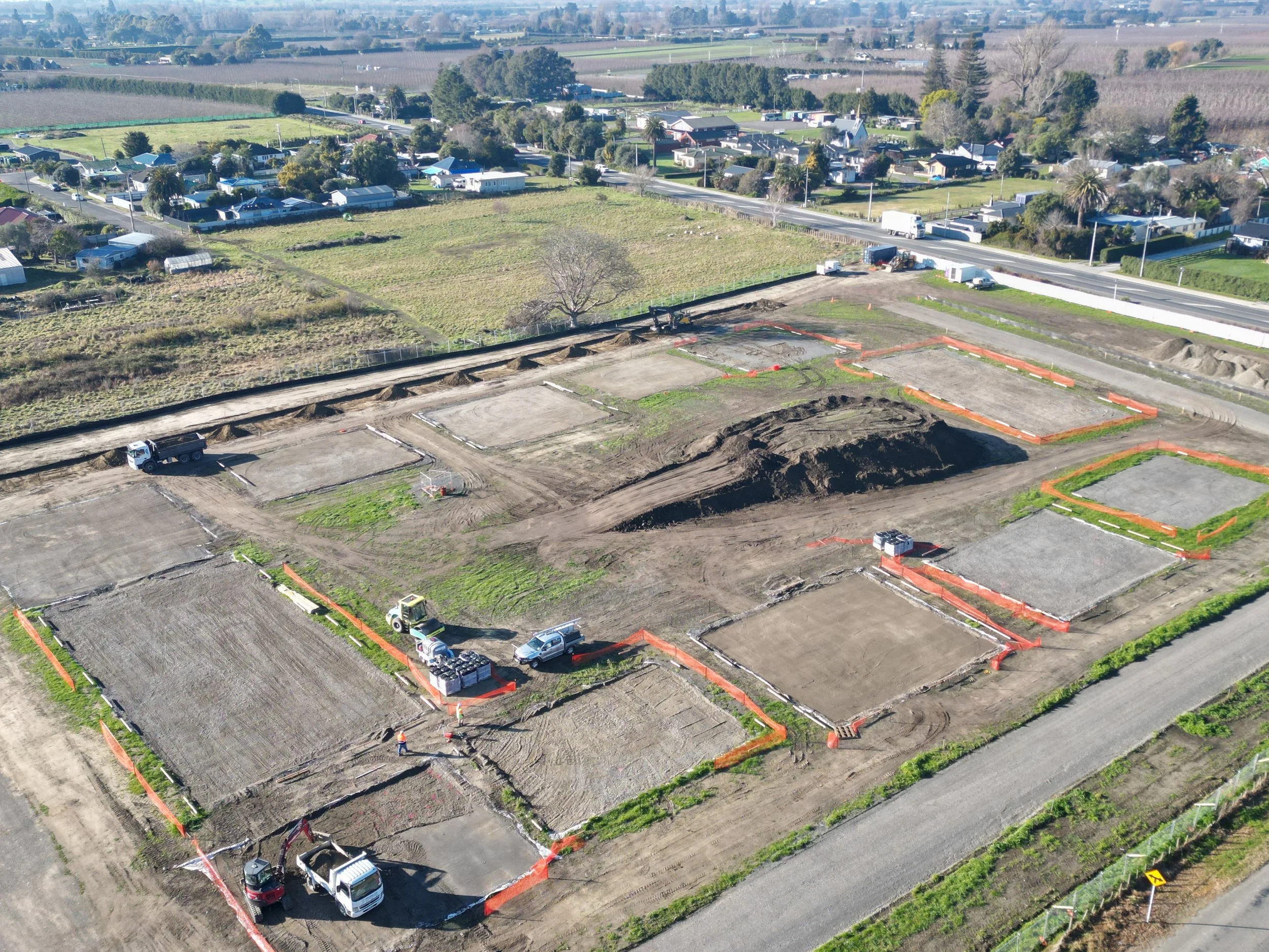 Aerial view of a construction site with marked plots and construction vehicles, surrounded by green fields and a nearby residential area.