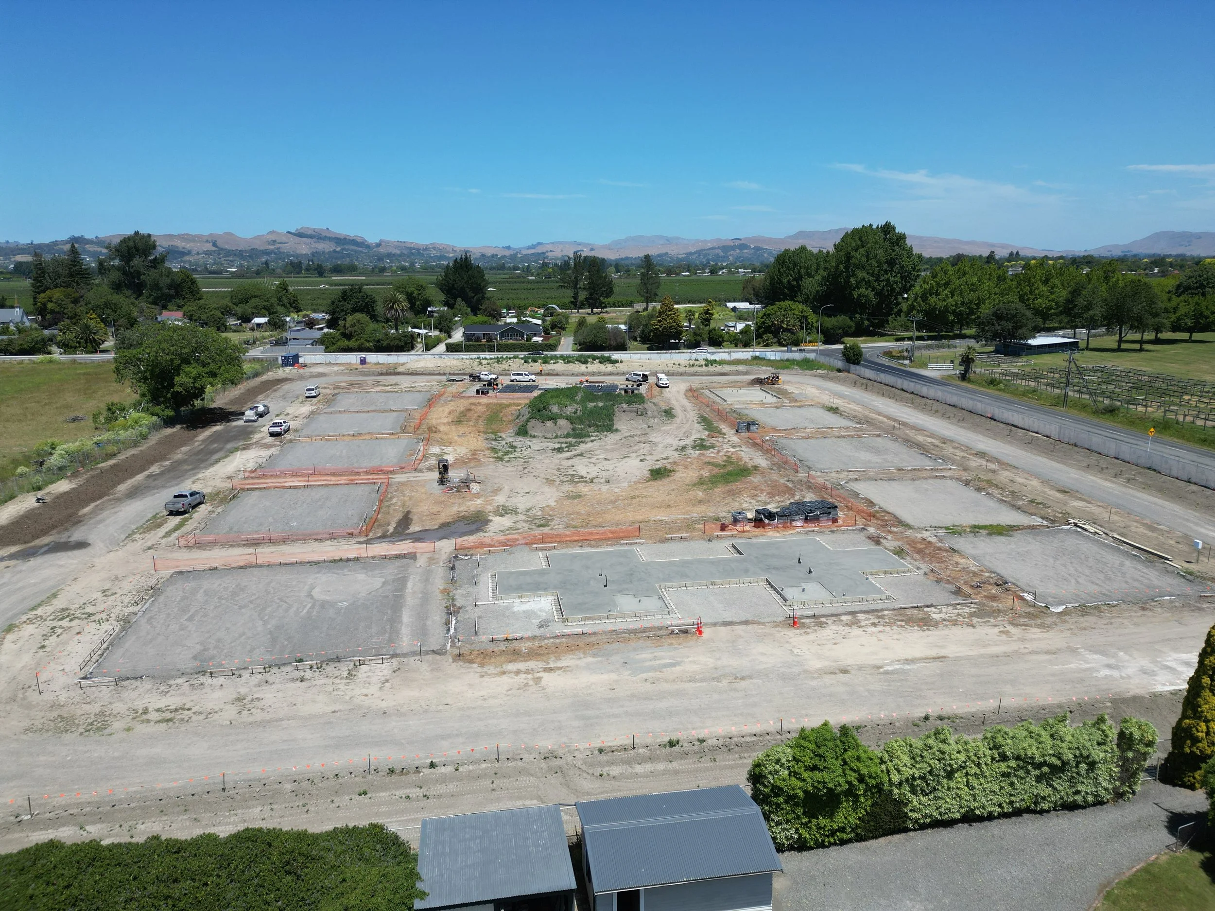 A construction site with multiple concrete foundations, surrounded by dirt and orange safety barriers. Several vehicles are parked nearby, and trees and fields are in the background under a clear blue sky.