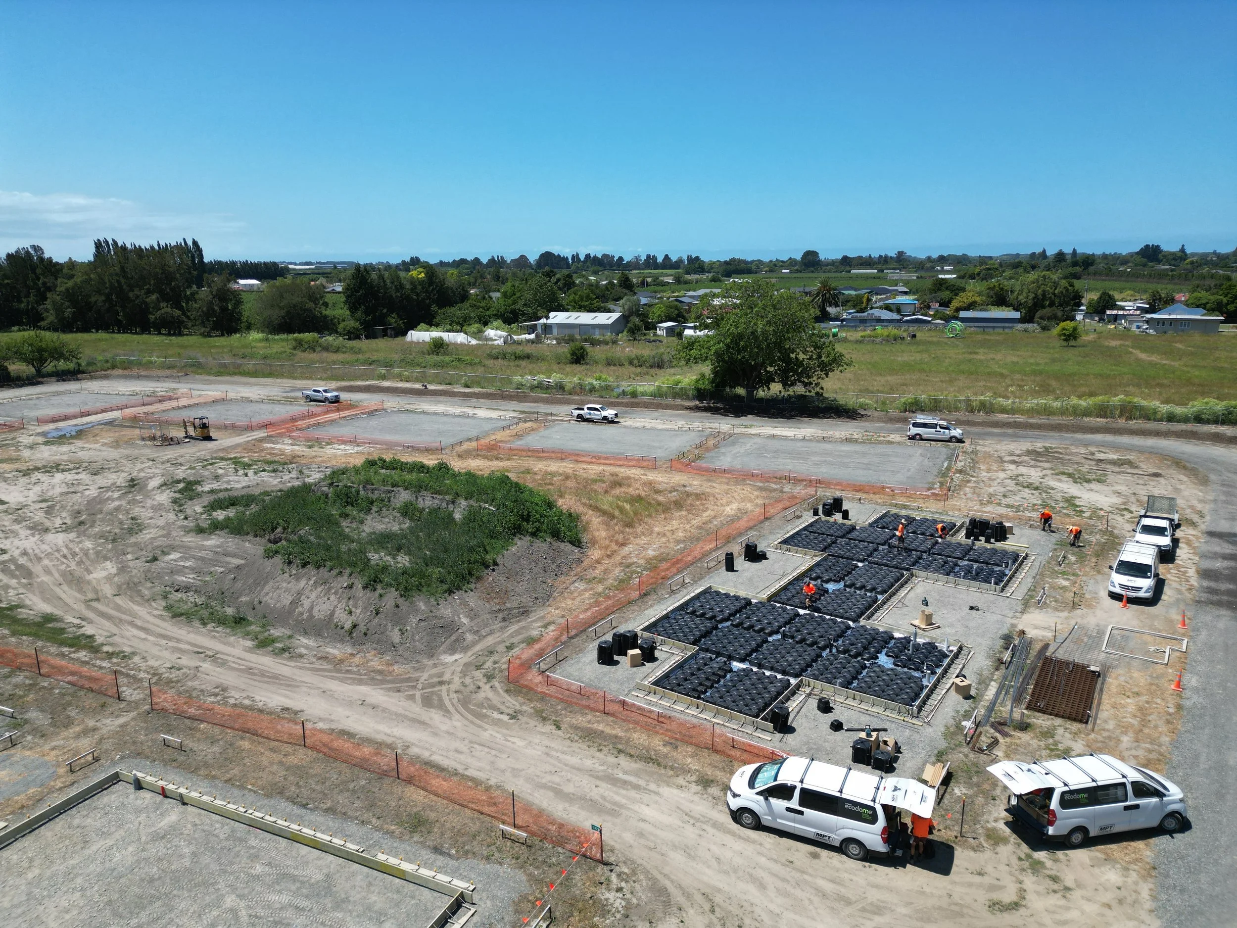 Construction site with vehicles, workers, and materials; fenced areas, groundwork preparation, and grassy surroundings.