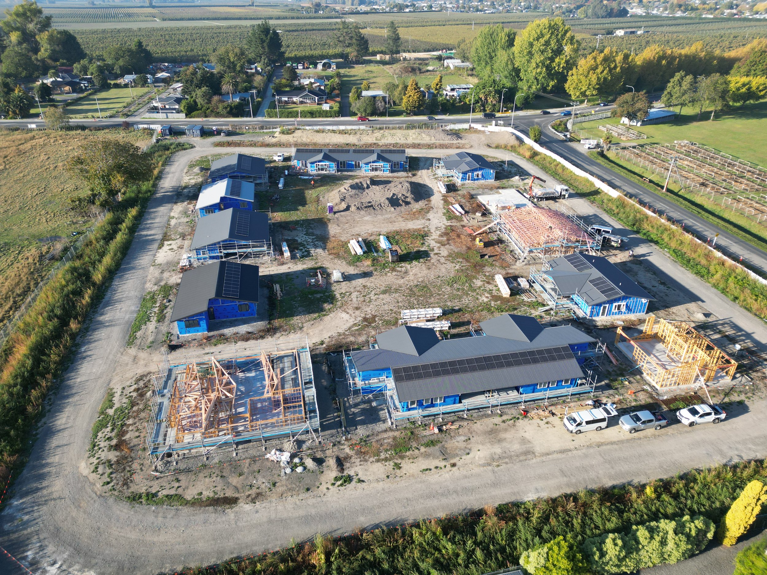 Aerial view of a construction site for residential buildings, featuring multiple houses in various stages of construction with wooden frames and blue tarps. The site is surrounded by roads and fields, with nearby residential areas and trees.