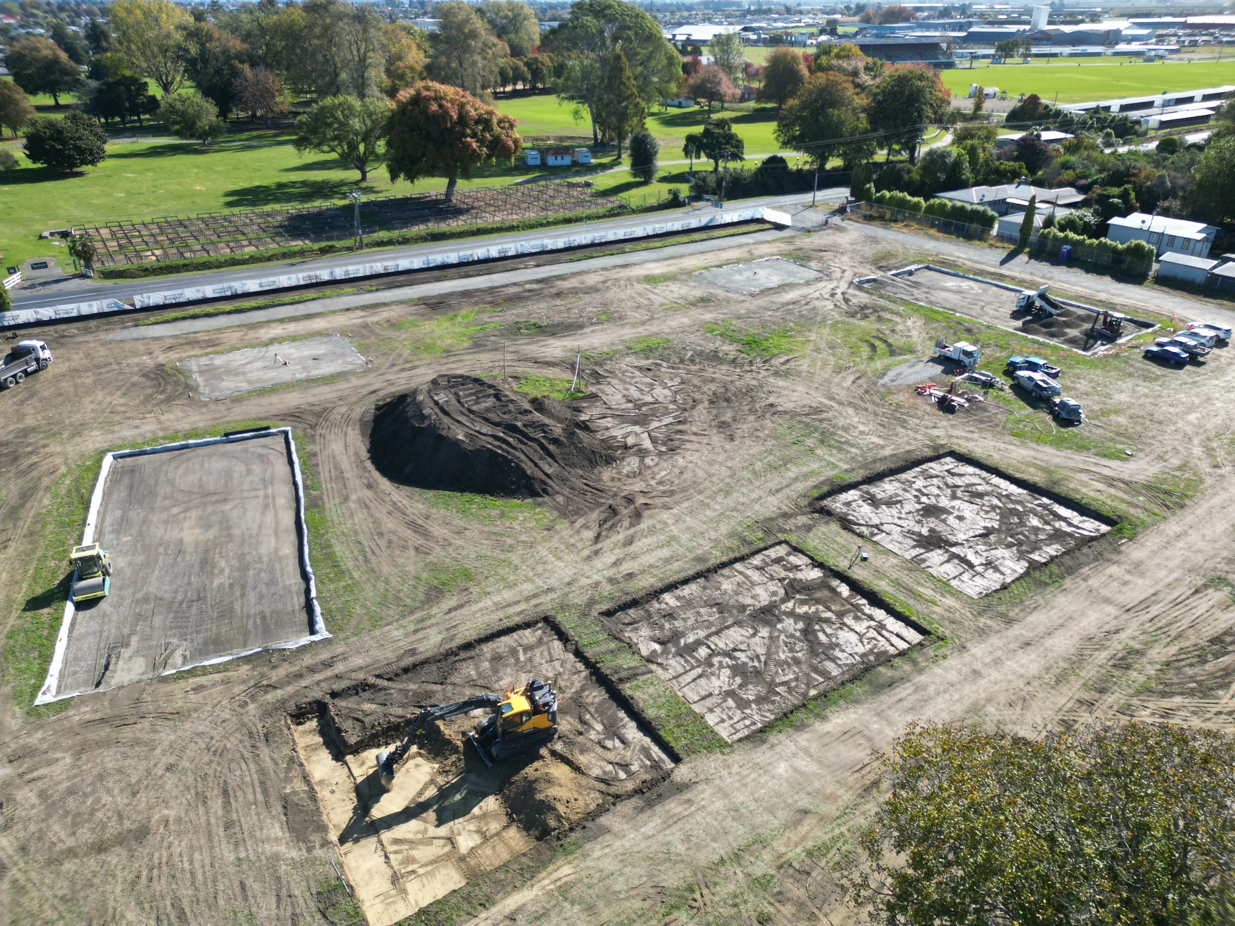Aerial view of a construction site with several excavated areas, piles of dirt, and construction vehicles, surrounded by greenery and roads.