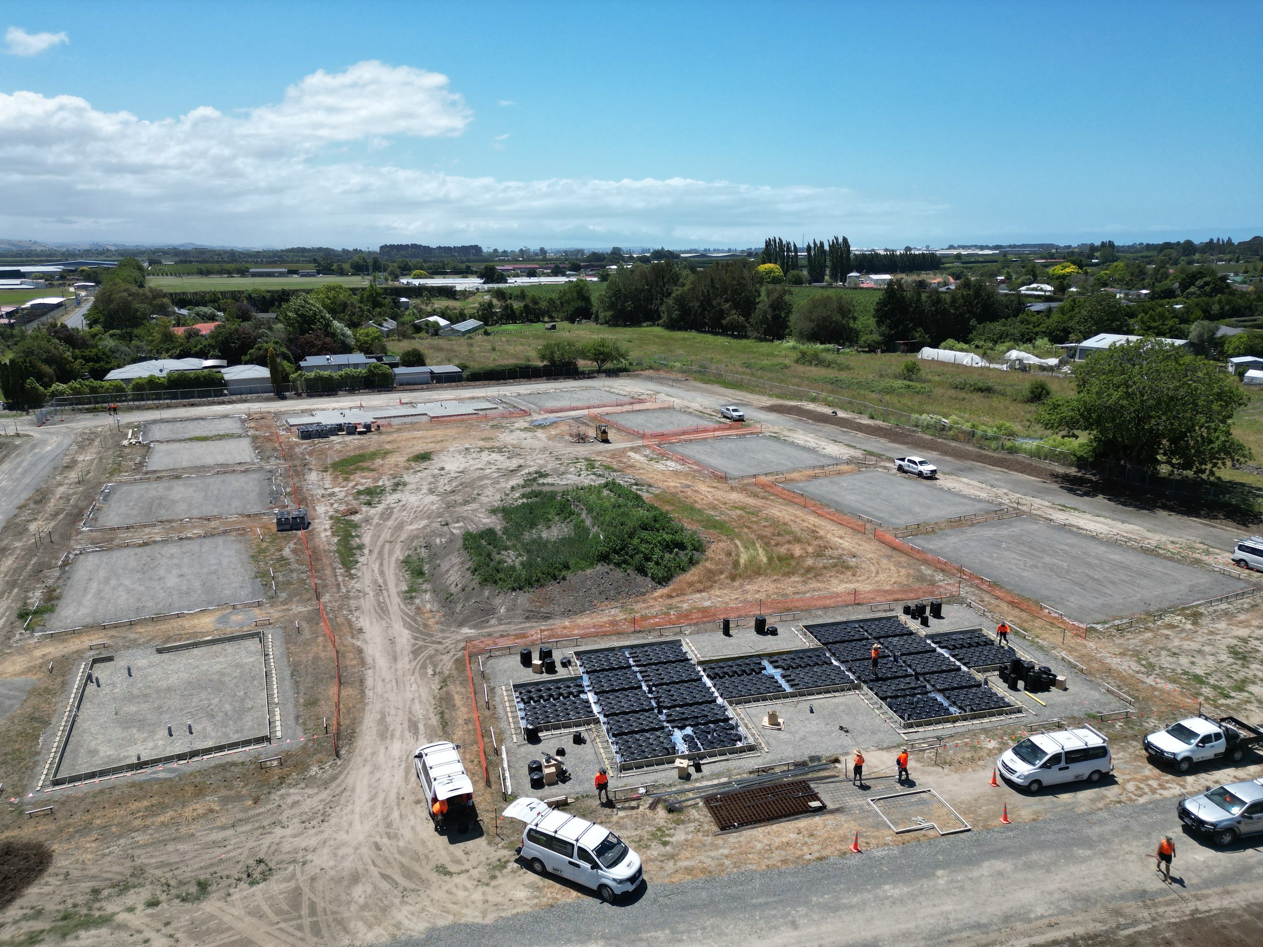 Aerial view of a construction site with several concrete foundations, surrounded by vehicles, workers in orange uniforms, and green fields in the background.