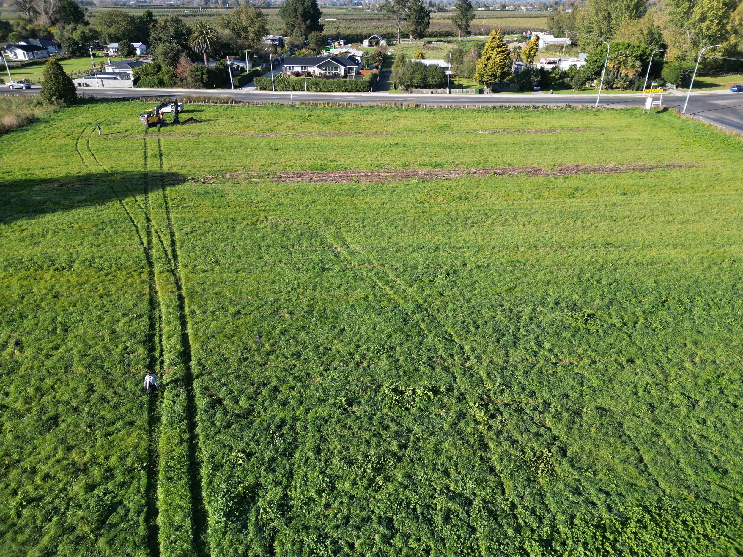 Aerial view of a large green field with tire tracks leading to an excavator near a road, surrounded by residential houses and trees.