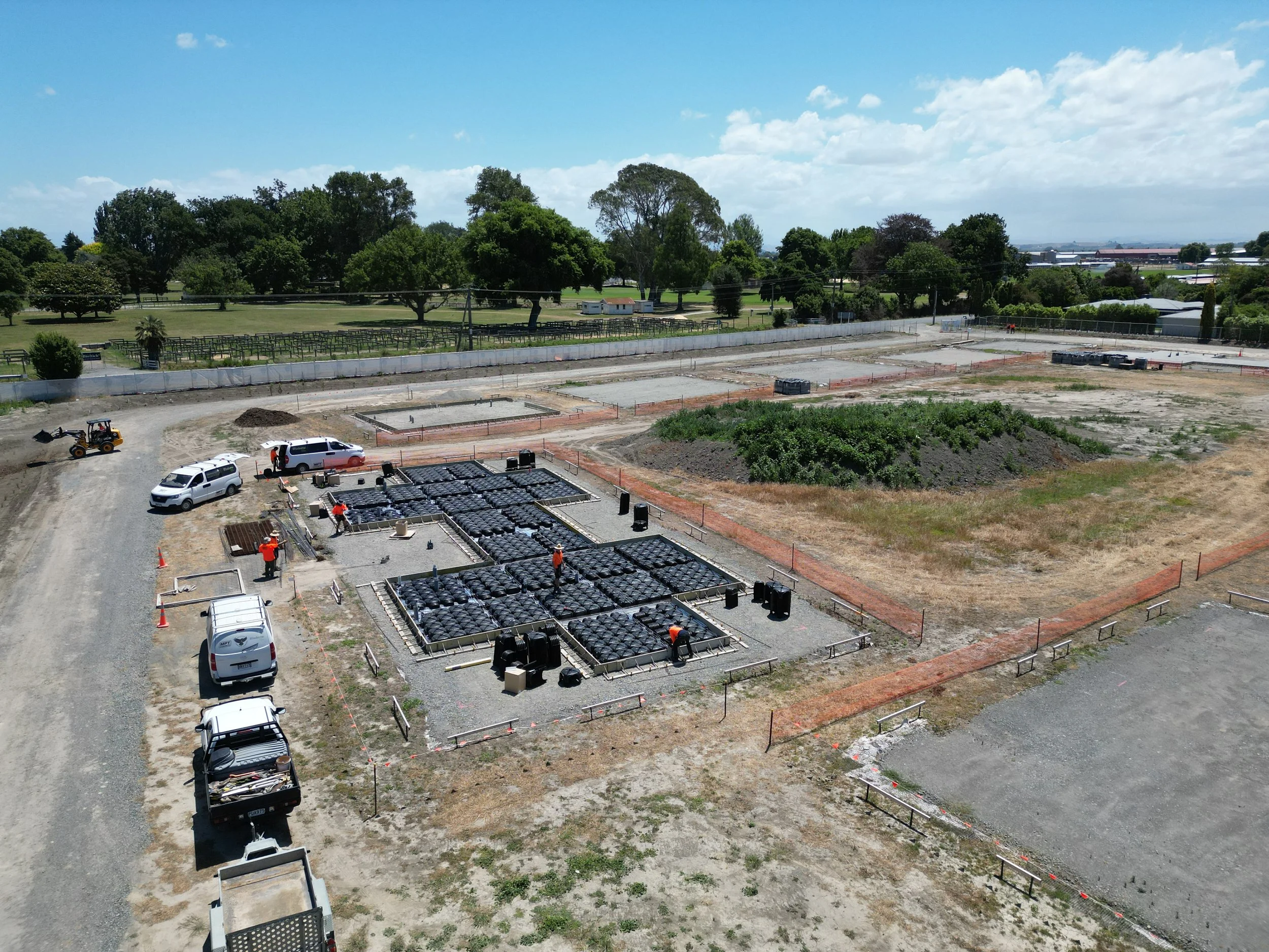 Aerial view of a construction site with workers, vehicles, and building materials. The site includes multiple sections with black grids and an open area surrounded by fencing. Trucks and construction equipment are present. Trees and an open landscape