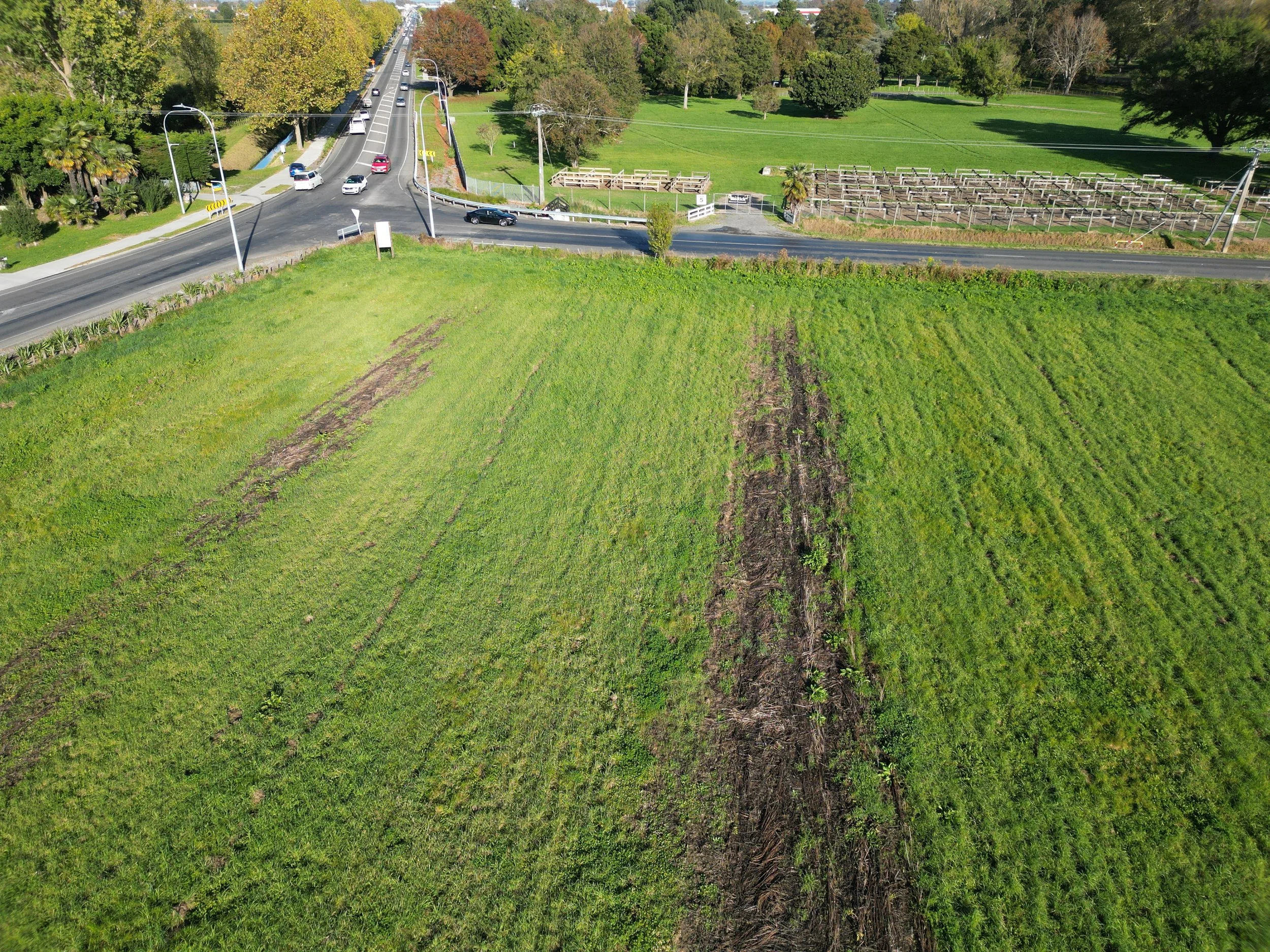 Aerial view of a grassy field with a road intersection and cars. The field has patches of brown soil and green grass. Trees and a fenced area are visible in the background.