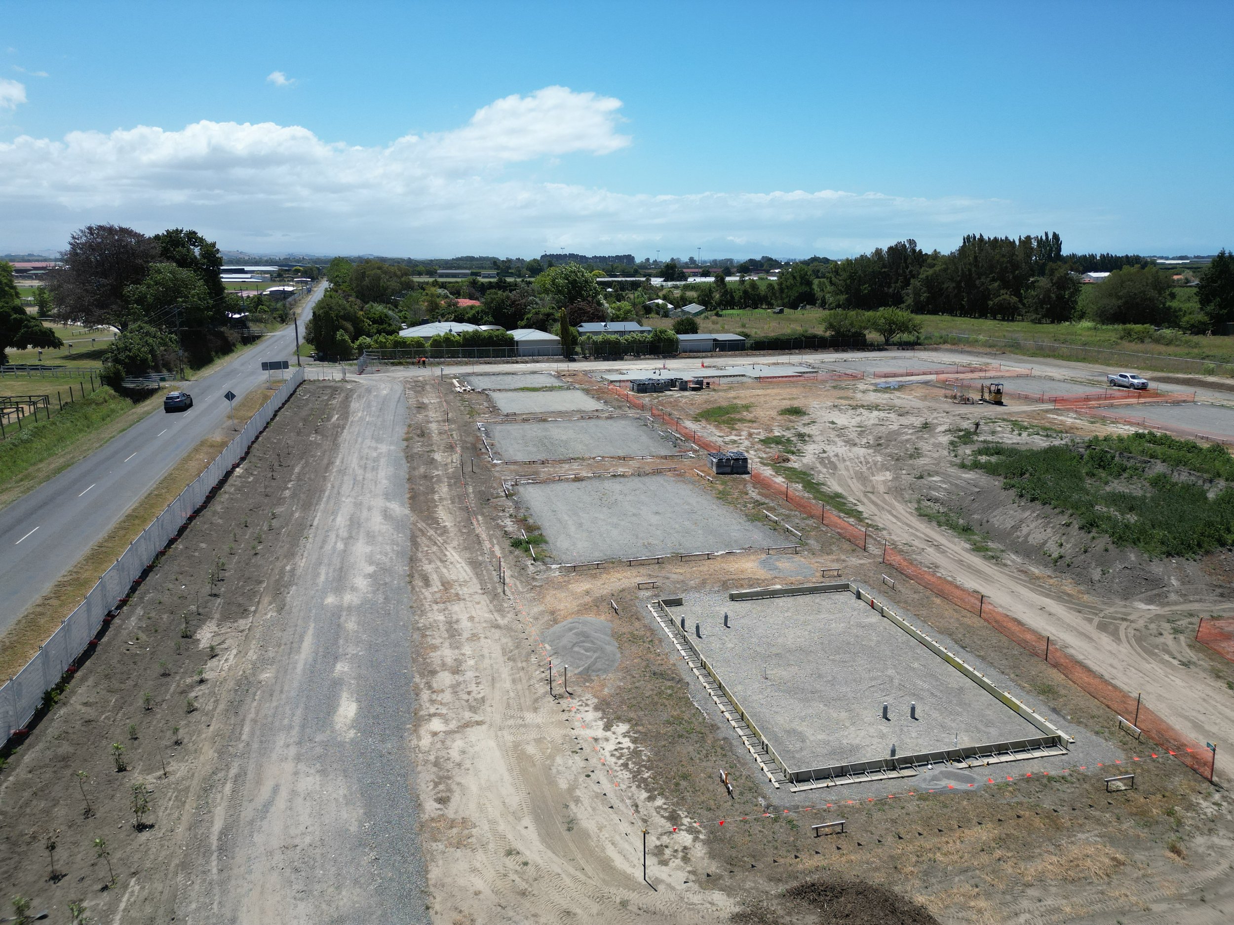 Aerial view of a construction site with several rectangular concrete foundations, surrounded by dirt and grass, adjacent to a road with a car and a fence line.
