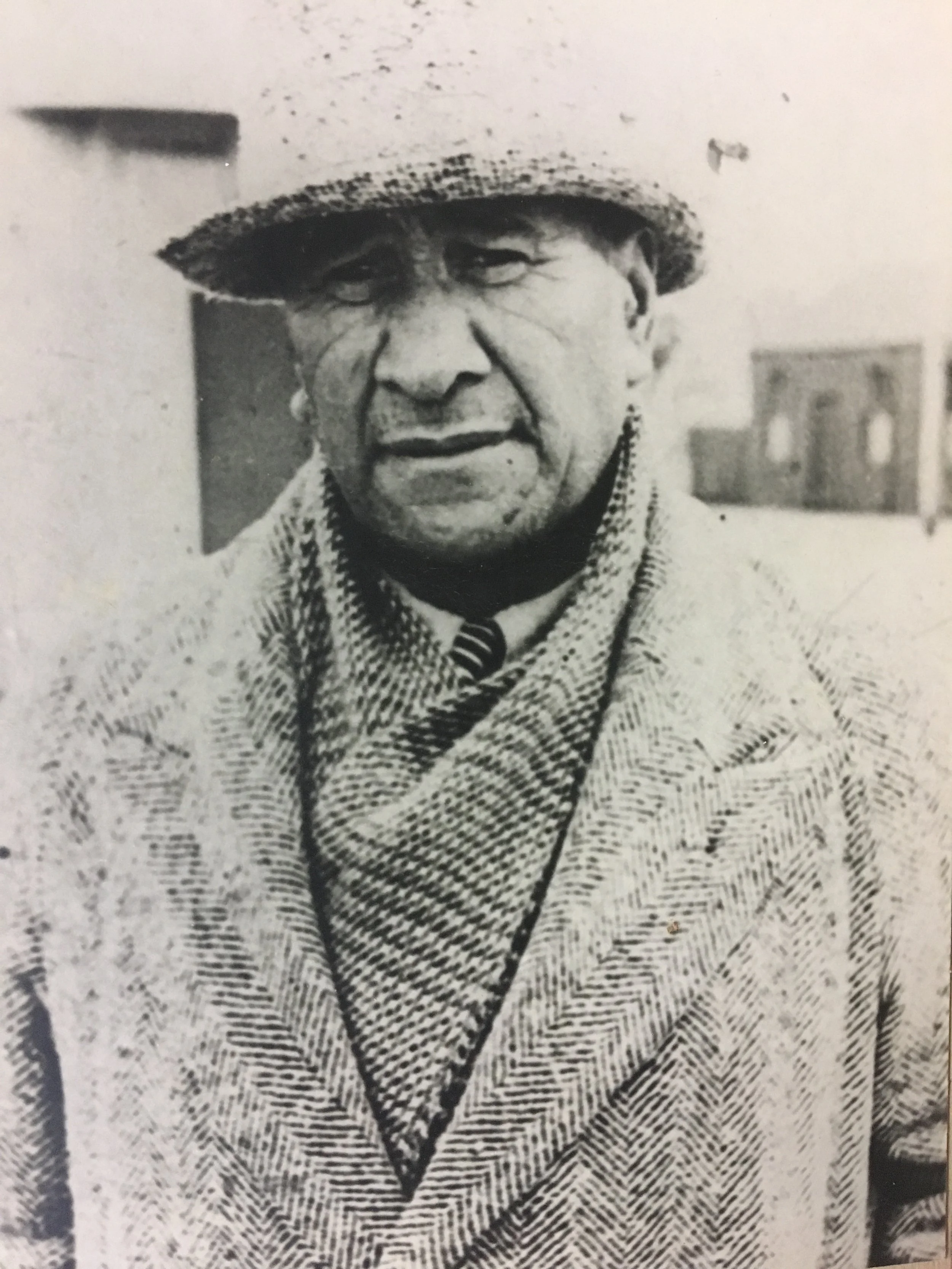 Black and white photo of a man wearing a hat and herringbone coat.
