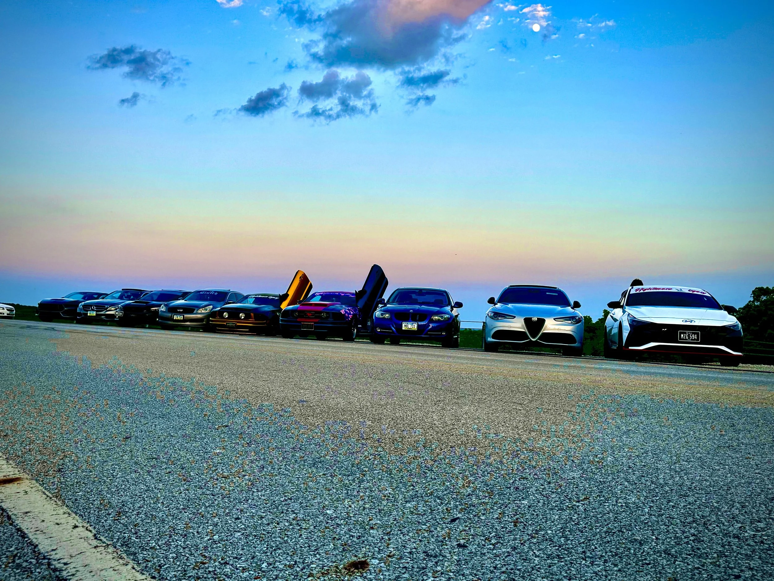 Row of sports cars parked on a road at sunset with a clear sky and scattered clouds.