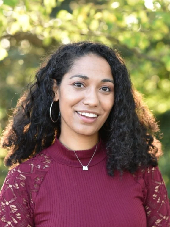 Smiling woman with curly black hair, hoop earrings, dressed in a maroon lace-sleeve top, outdoors with green foliage background.