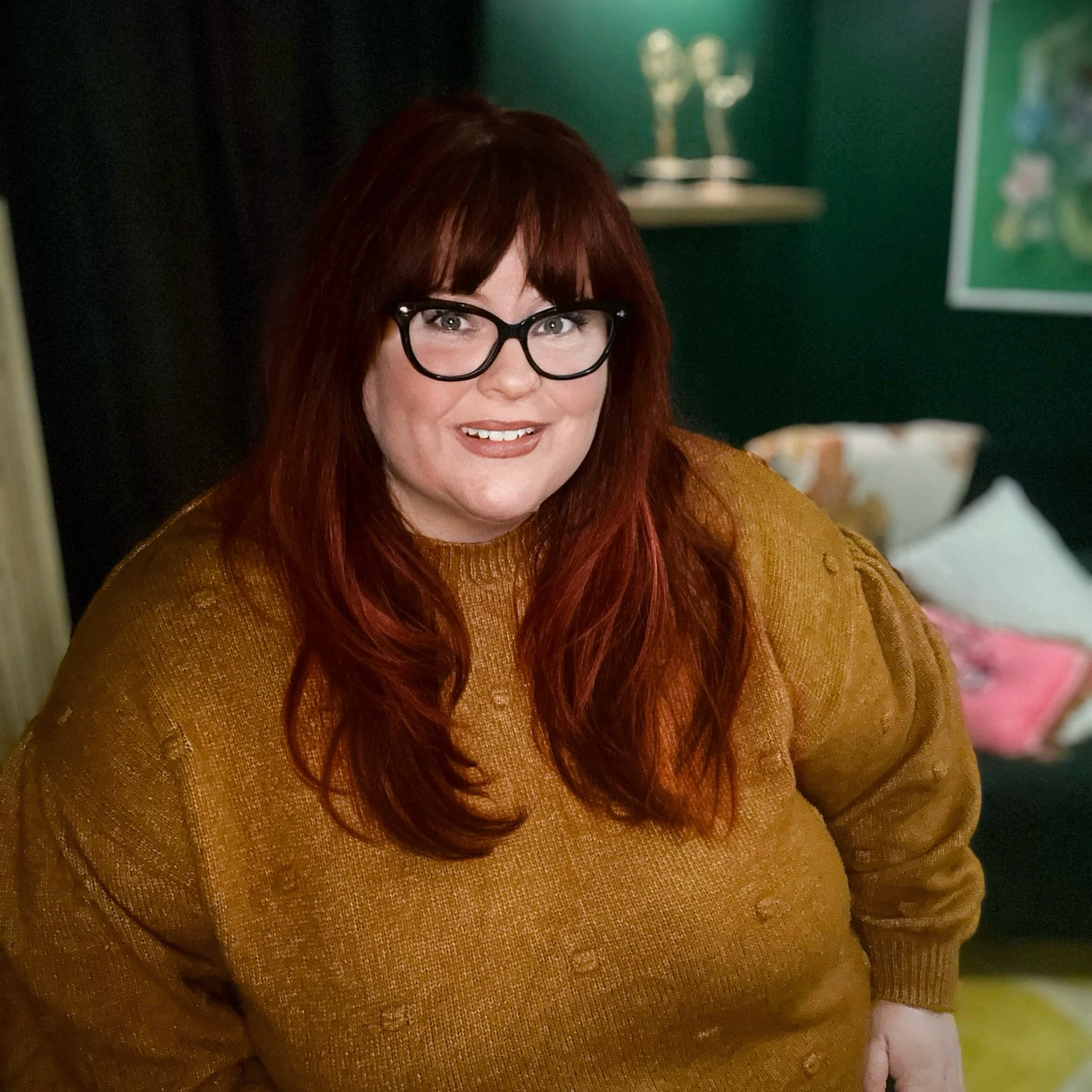 A woman with long red hair, glasses, and a mustard sweater smiling in a cozy, decorated room.