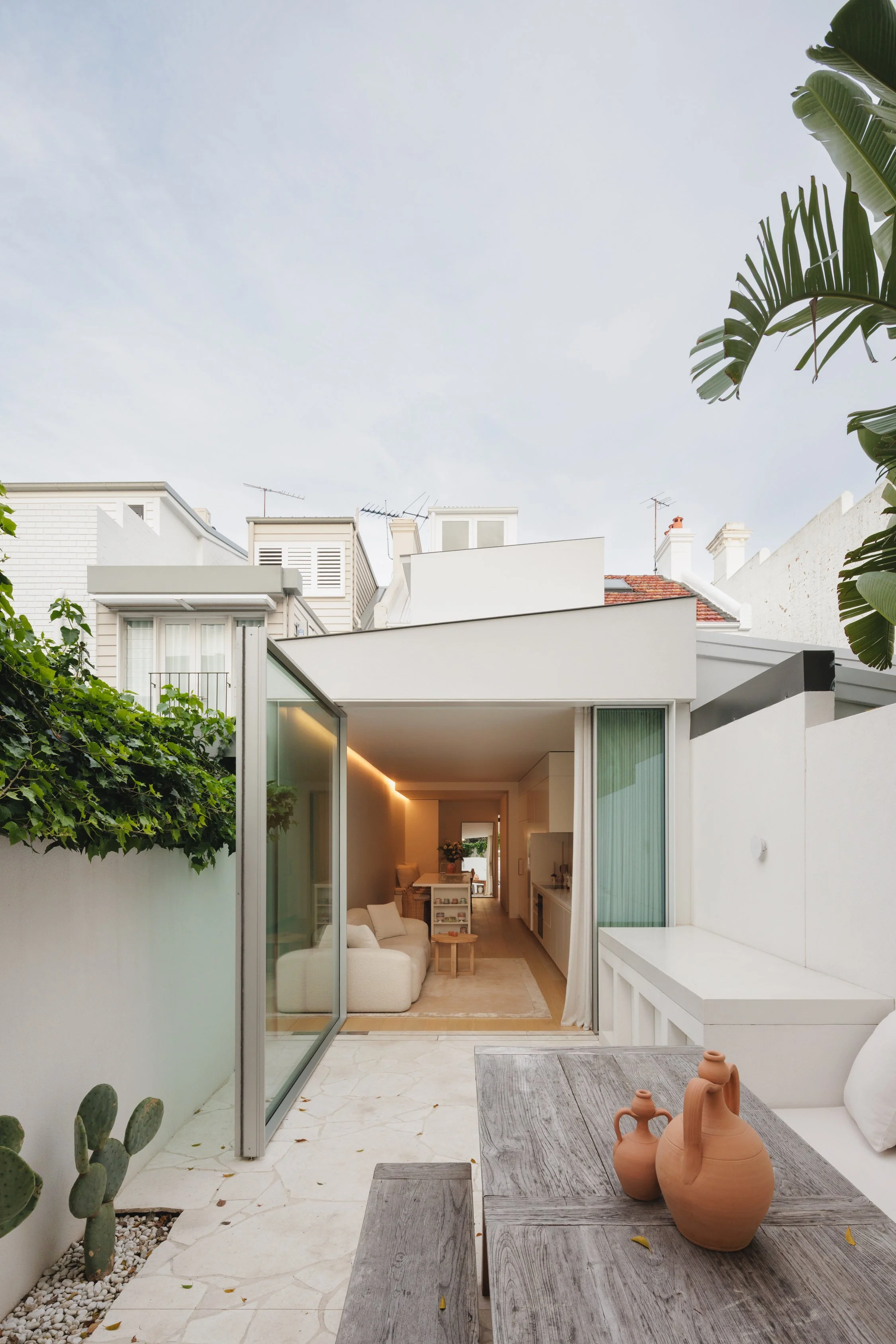 Modern open-plan outdoor patio with a view into a minimalist living room, featuring a wooden table with terracotta vases, white walls, and lush greenery around.