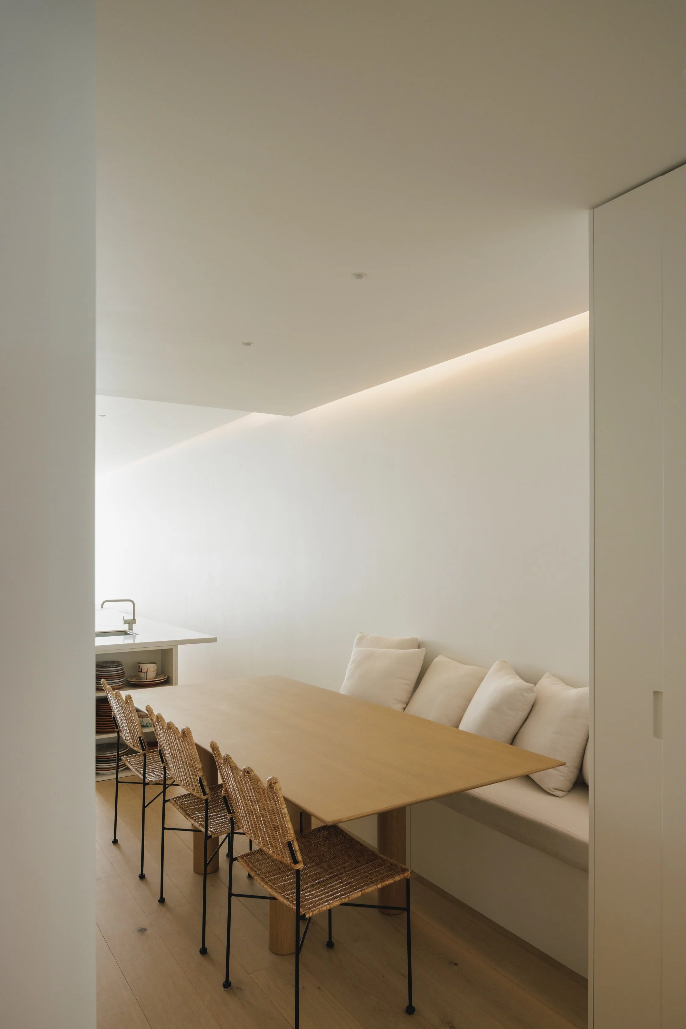 Minimalist dining area with a timber table, wicker chairs, built-in banquette seating and soft lighting.