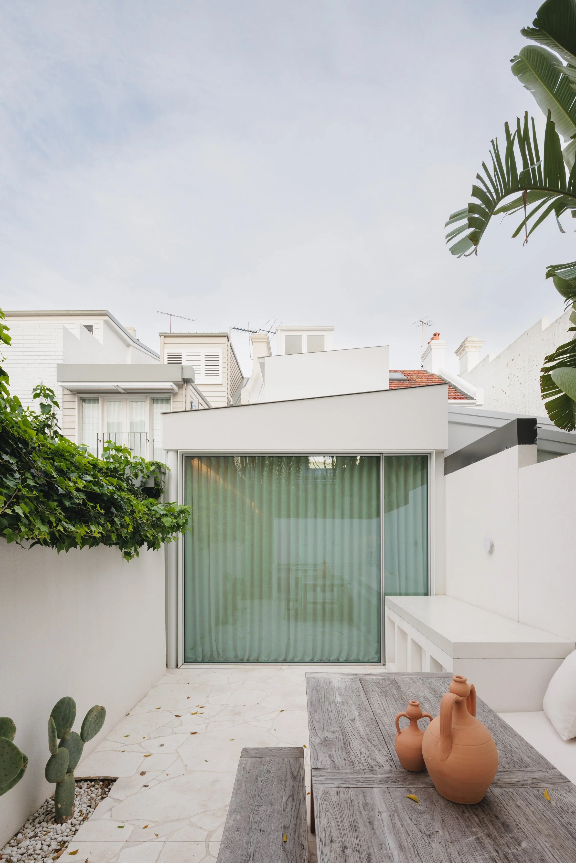 Outdoor patio area with a wooden table holding terracotta vases, white walls, and a glass sliding door with green curtains. Surrounding greenery includes cacti and leafy plants.
