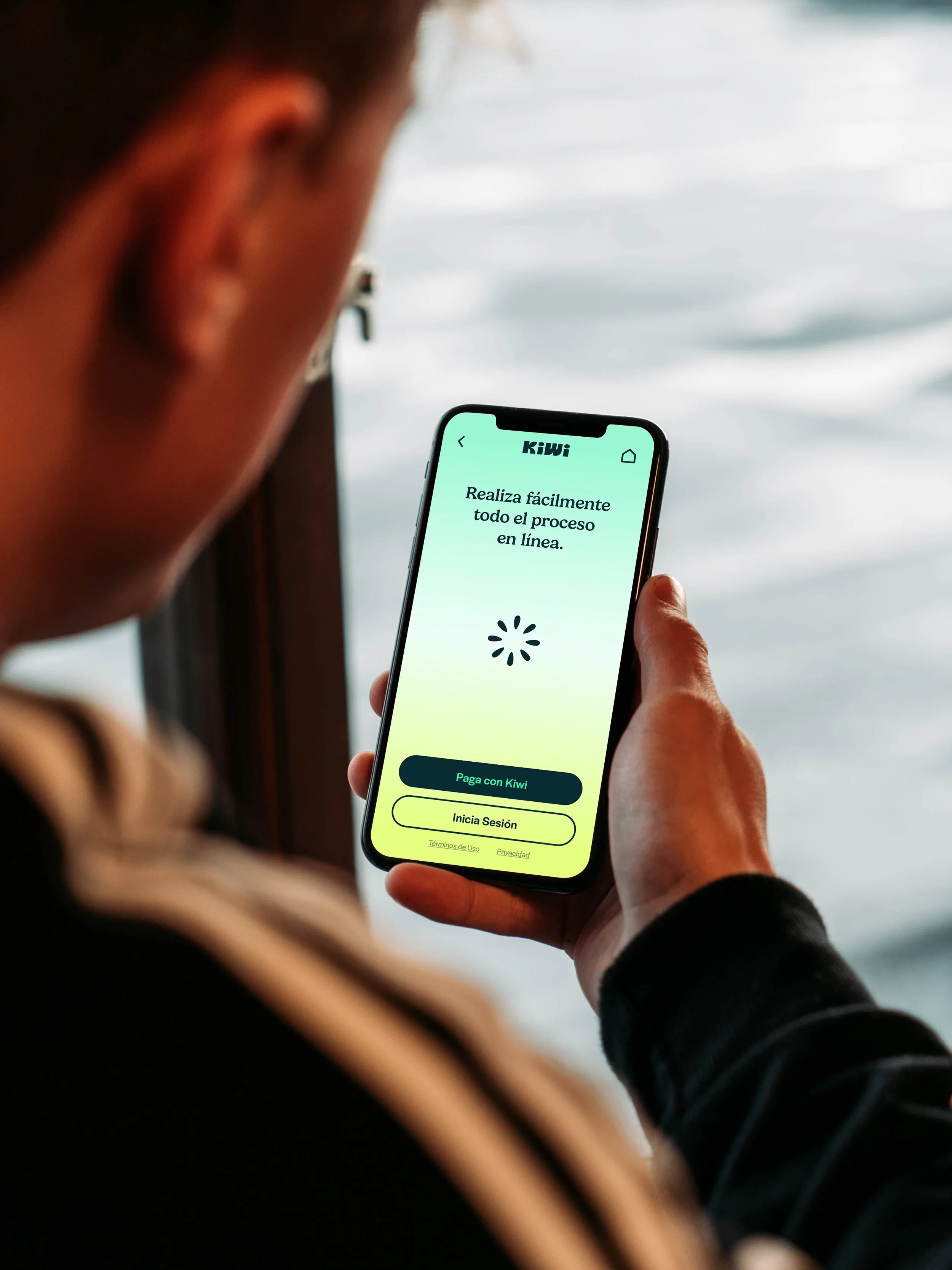 Persona sosteniendo un teléfono móvil con la pantalla de una aplicación llamada Kiwi, mostrando opciones para realizar pagos y sesiones en línea, con fondo de agua.
