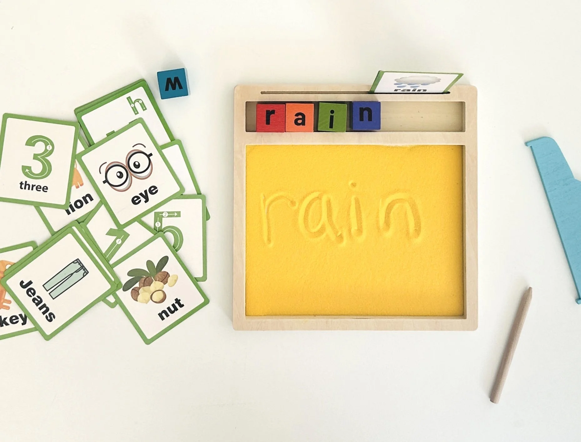 A child's wooden spelling and picture activity set showing the word 'rain' written in sand on a yellow tray, with letter tiles spelling 'rain' above it, and picture flashcards with words like 'three,' 'eye,' 'nut,' and 'jeans' scattered on a white surface.
