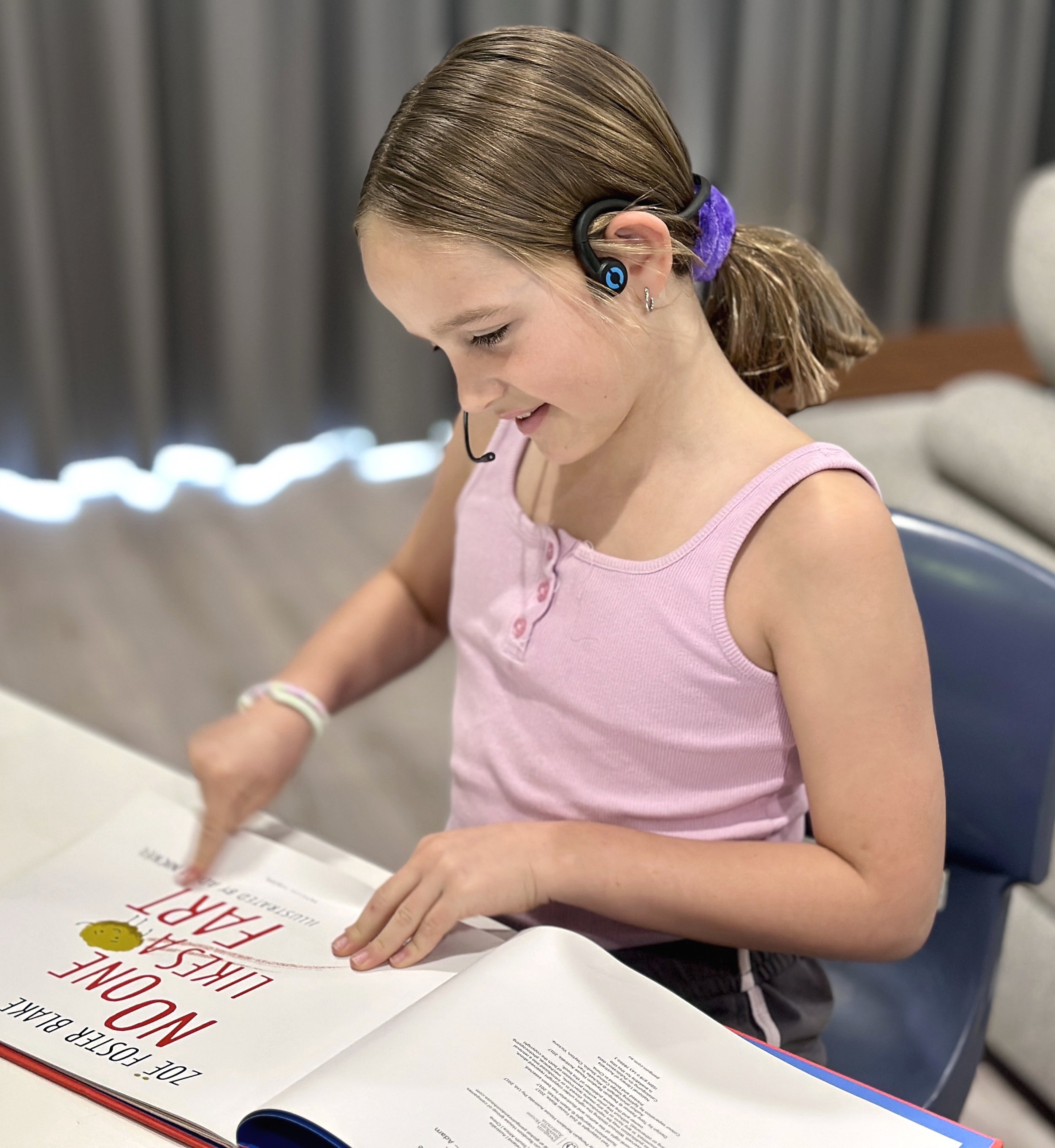 Young girl with brown hair in a ponytail, wearing a pink sleeveless top and a headset, smiling as she reads a book titled 'No One Likes a Fart'.