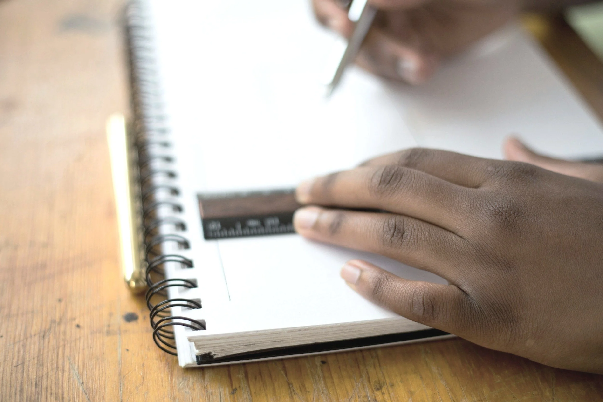 A student is drawing a writing graphic organiser with a black ruler on a white spiral notebook, with a pen on a wooden desk in the background.