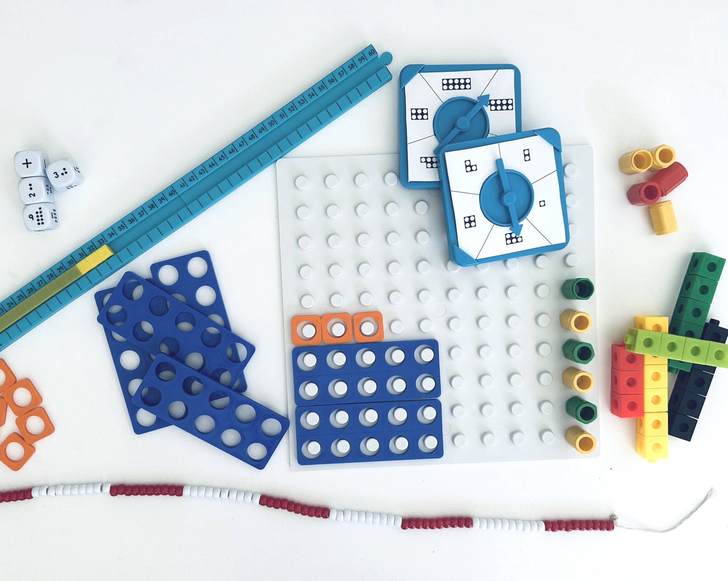 Table with various colourful math manipulatives, including dice, spinners, and counters, some arranged on a white pegboard, with two blue Numicon shapes and a blue cube spinner, and a red-and-white beaded number line at the bottom.