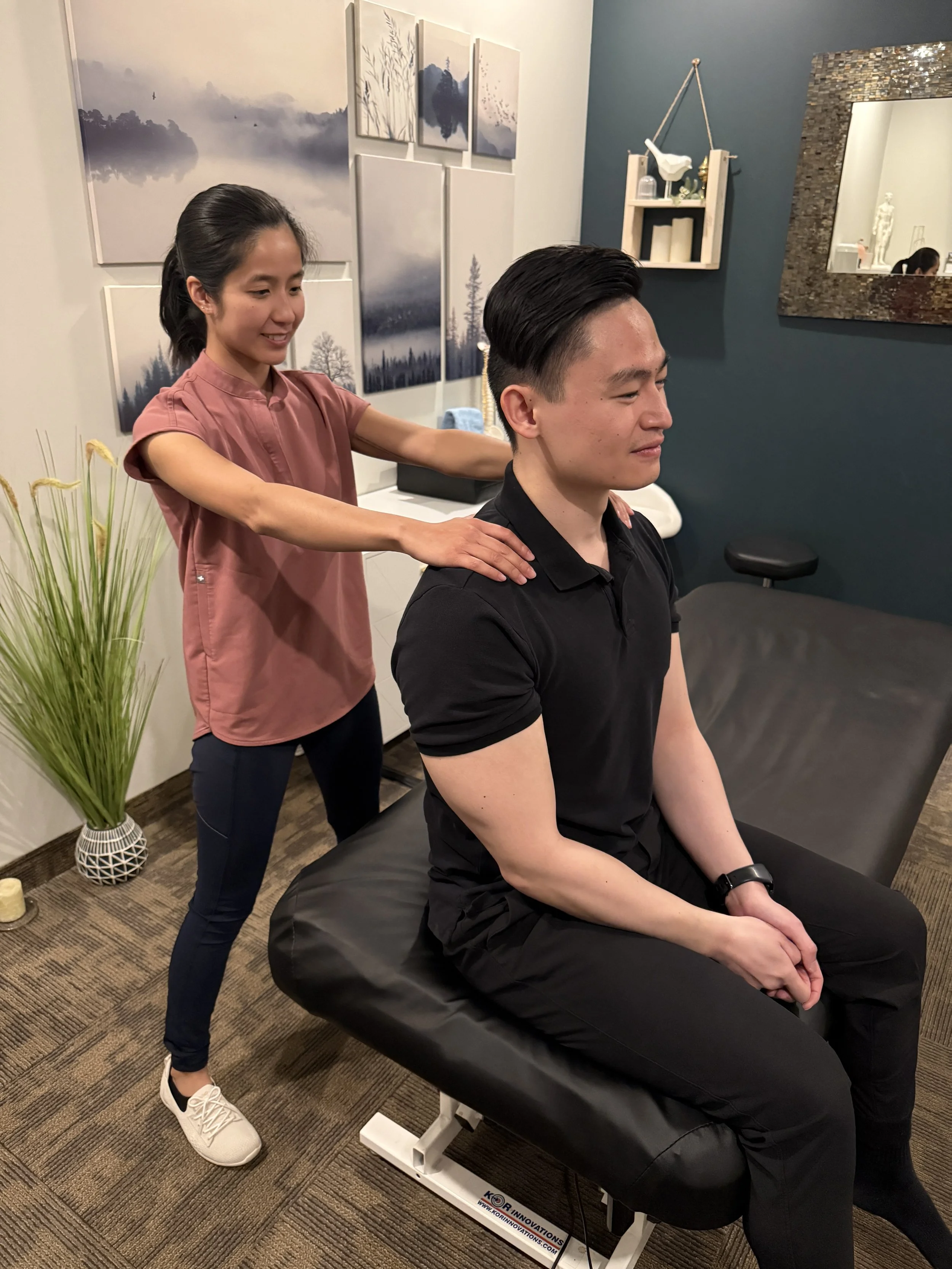 A woman is giving a shoulder massage to a man sitting on a massage table in a room decorated with landscape artwork, a mirror, and shelves with decorative items. The woman is smiling and wearing a pink shirt, while the man is wearing a black shirt and appears relaxed.