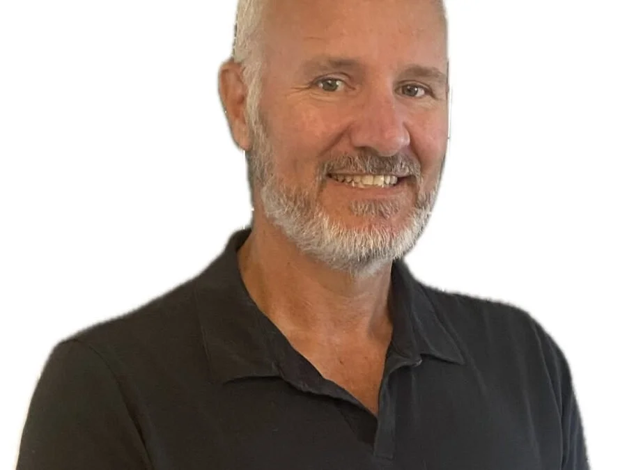 A middle-aged man with a short white beard and light eyes, smiling, wearing a black collared shirt against a white background.