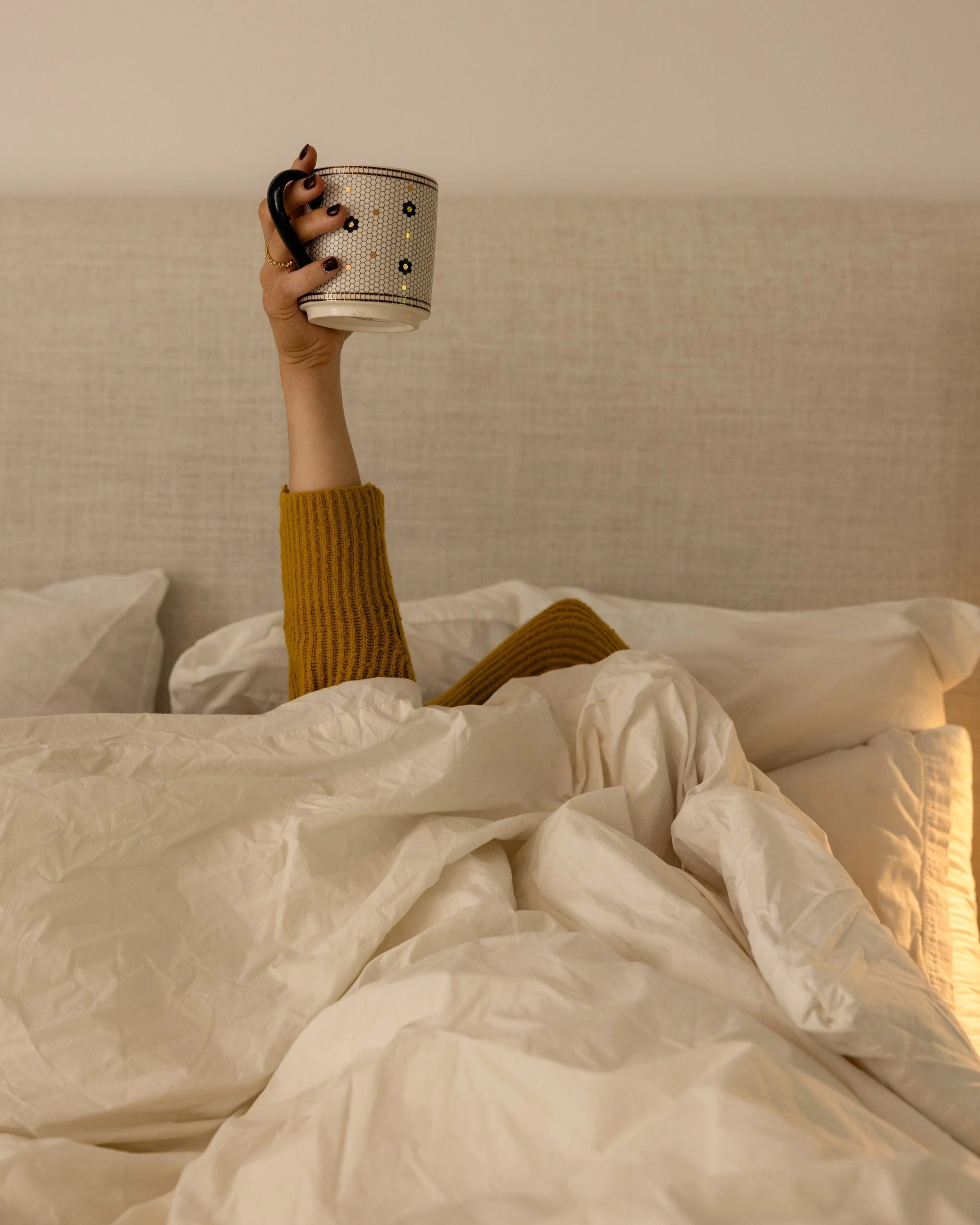 Lauren Harvey- ATL Sleep consultant lying in bed with a blanket over her head, holding up a coffee mug — capturing the exhaustion parents feel before sleep training