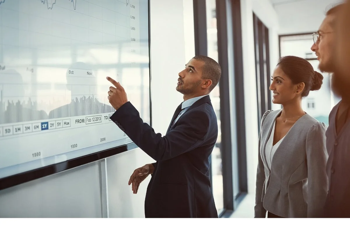 A man in a suit explains data on a large screen to three colleagues.