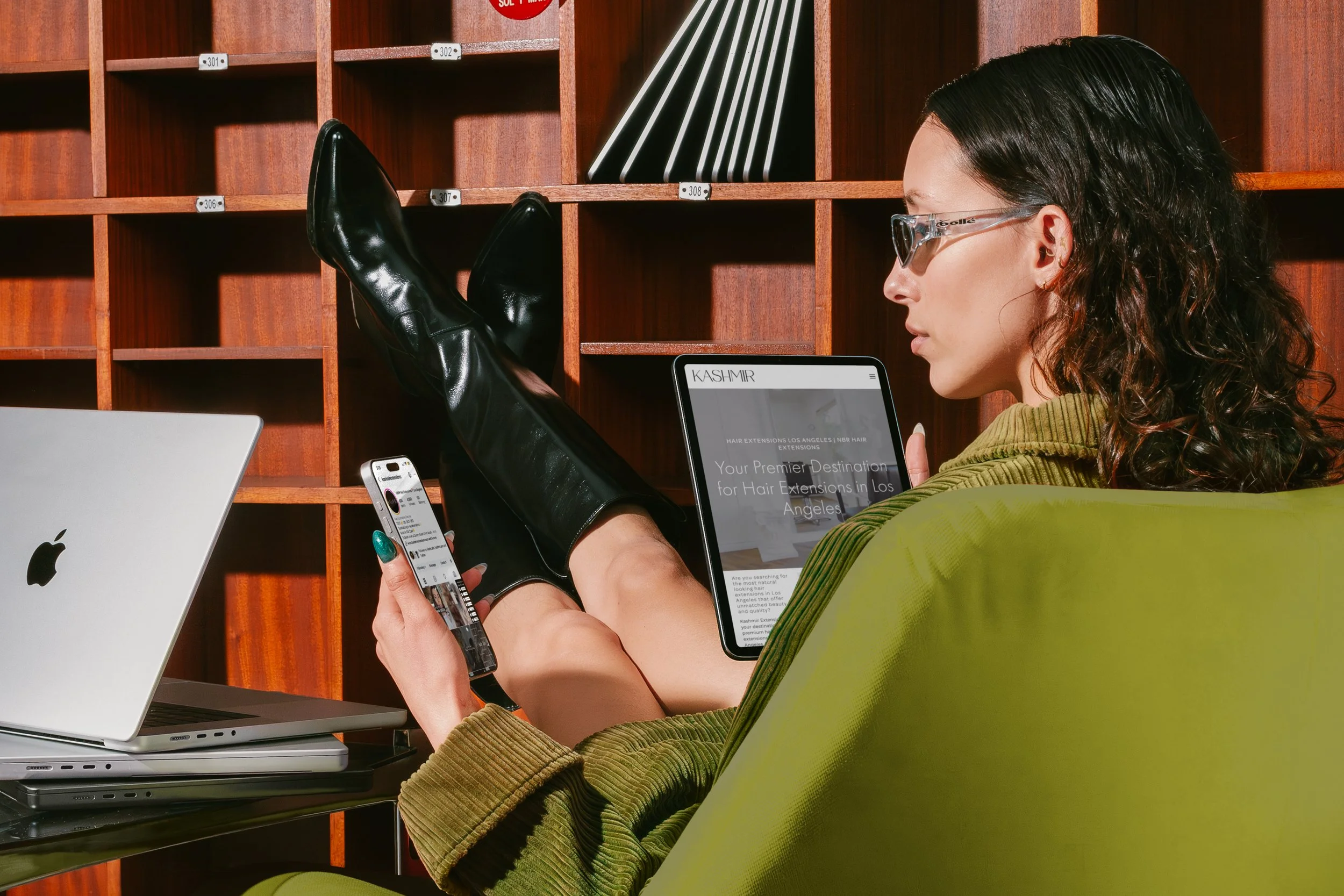 A woman wearing glasses and a green jacket is sitting with her legs up on a desk, using a smartphone and surrounded by multiple laptops, in a room with wooden wall cubbies and styled books.