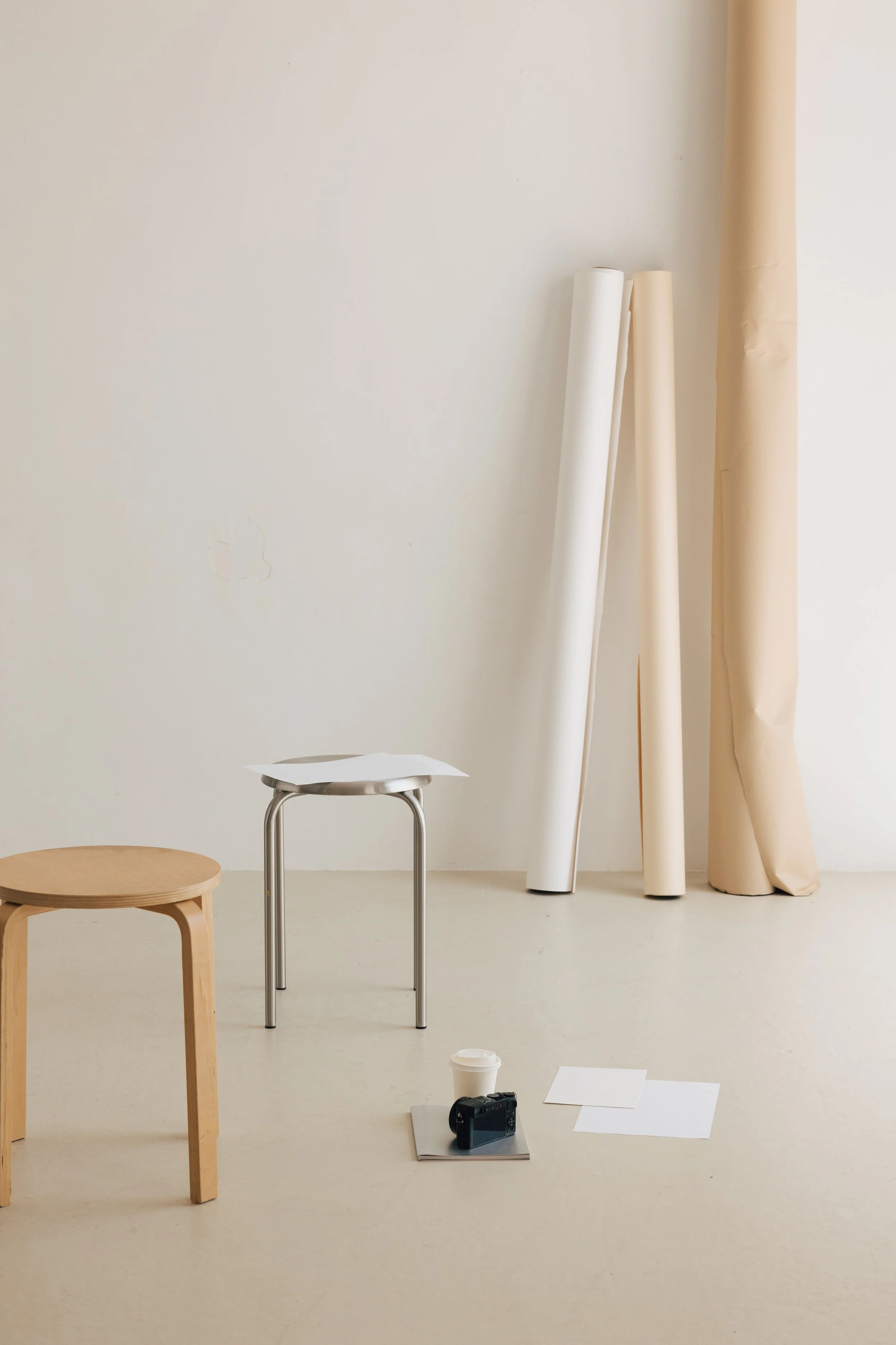 Minimalist indoor scene with a small wooden stool, a metal table with a sheet of paper on top, a camera, a white paper cup, and artist's rolls of paper or fabric in neutral tones against a plain light wall.