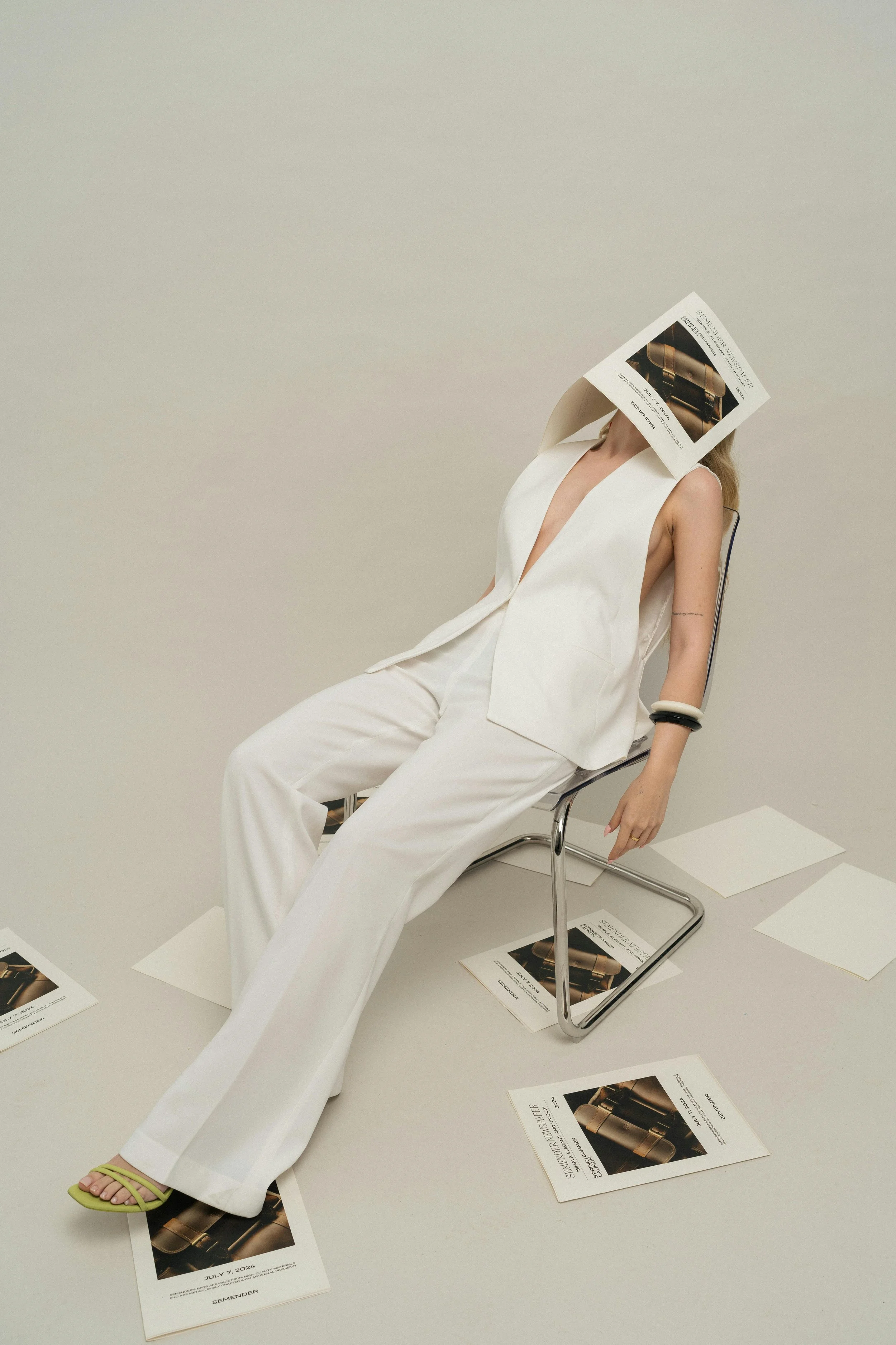 A woman dressed in a white sleeveless suit sitting on a chair with a magazine covering her face. Photographs of a product are on the floor and in her hand, with reproductions of the magazines spread around her.