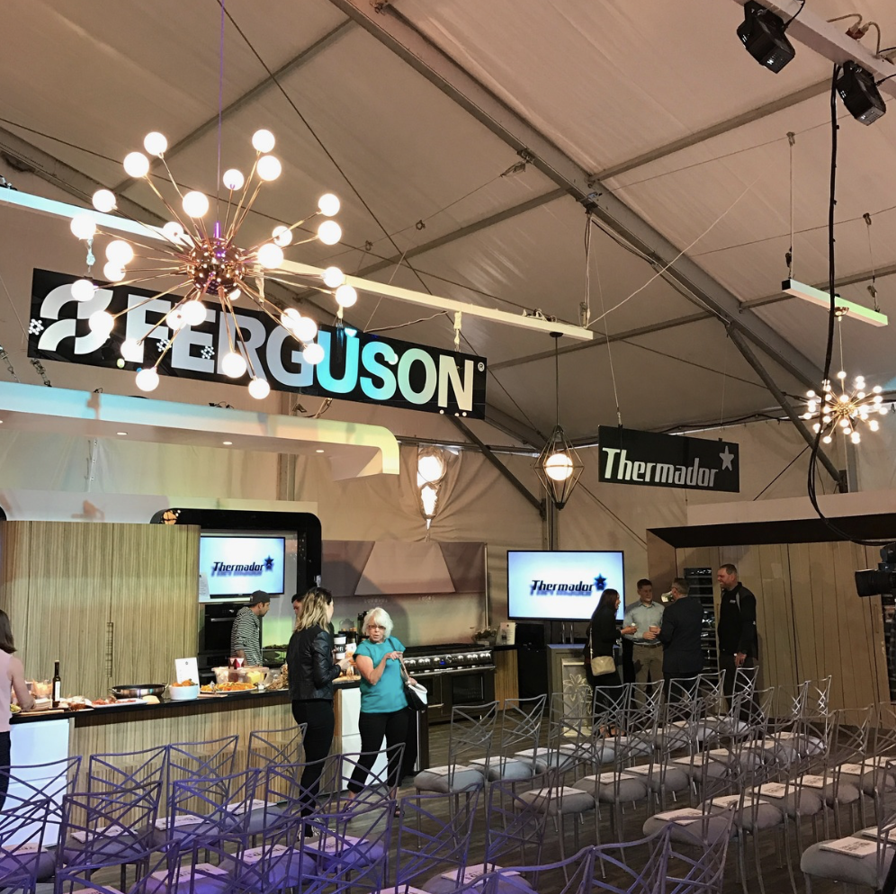 Interior of an event space with chairs, chandeliers, and a stage area displaying the FergUSON and Thermador signs, with a few people preparing and chatting.