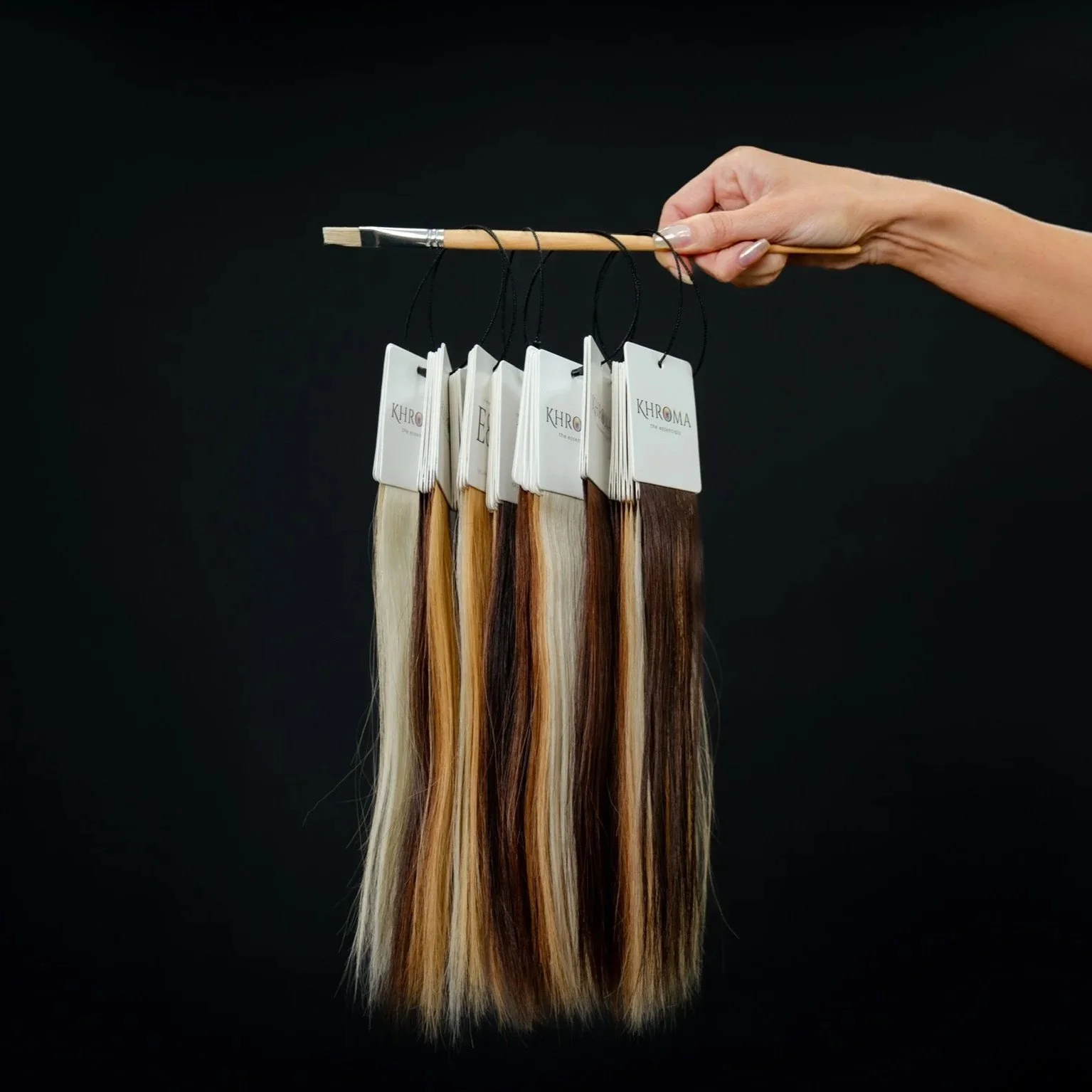 Hand holding a paintbrush with five hair color sample strips hanging from it, each labeled 'KHROMA' with various shades of blonde and brown hair.