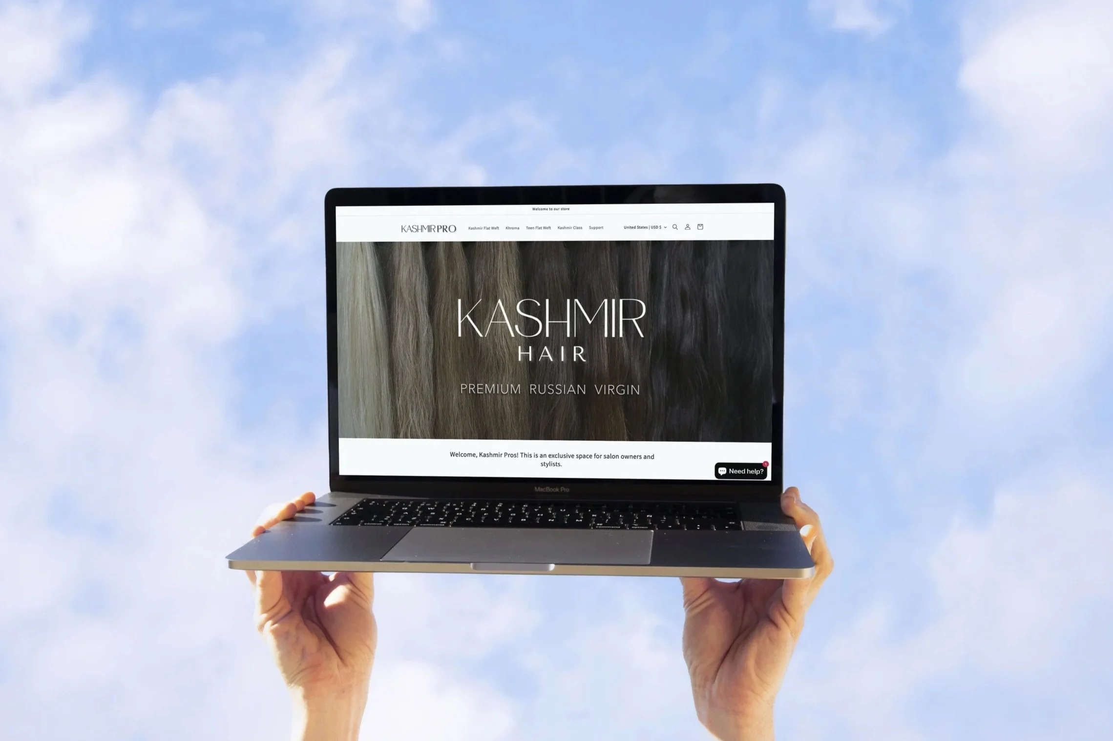 A person holding an open MacBook Pro laptop with a website about Kashmir hair displayed, against a backdrop of blue sky with scattered clouds.