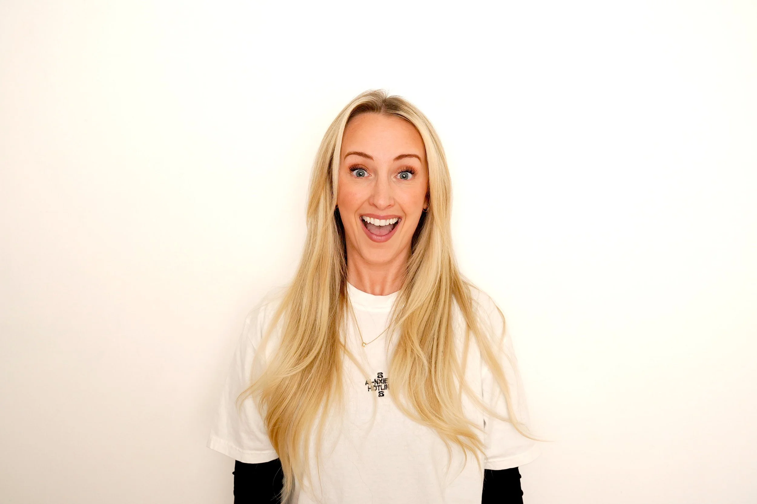 A woman with long blonde hair, wearing a white T-shirt with the phrase 'SAXY AND INDEPENDENT' written on it, smiling and standing against a plain white background.
