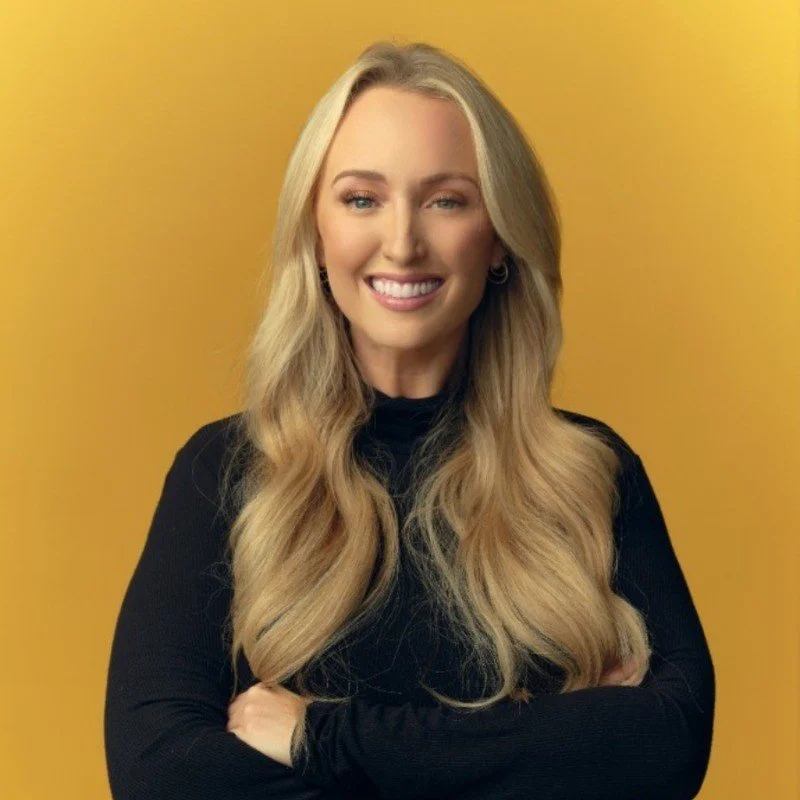 A woman with long blonde hair, smiling with arms crossed, standing against a yellow background.