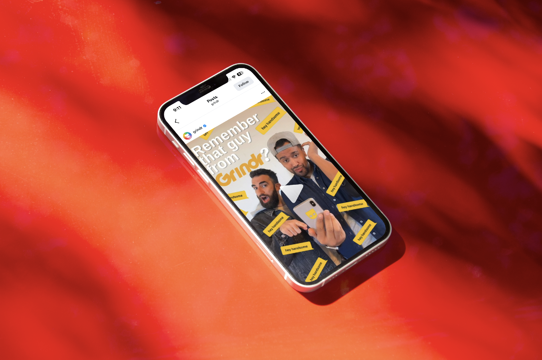 An iPhone displays a video thumbnail featuring two men taking a selfie, with text reading "Remember that guy from Grindr?" and yellow stickers with "hey handsome" written on them surrounding the men, on a red and purple abstract background.