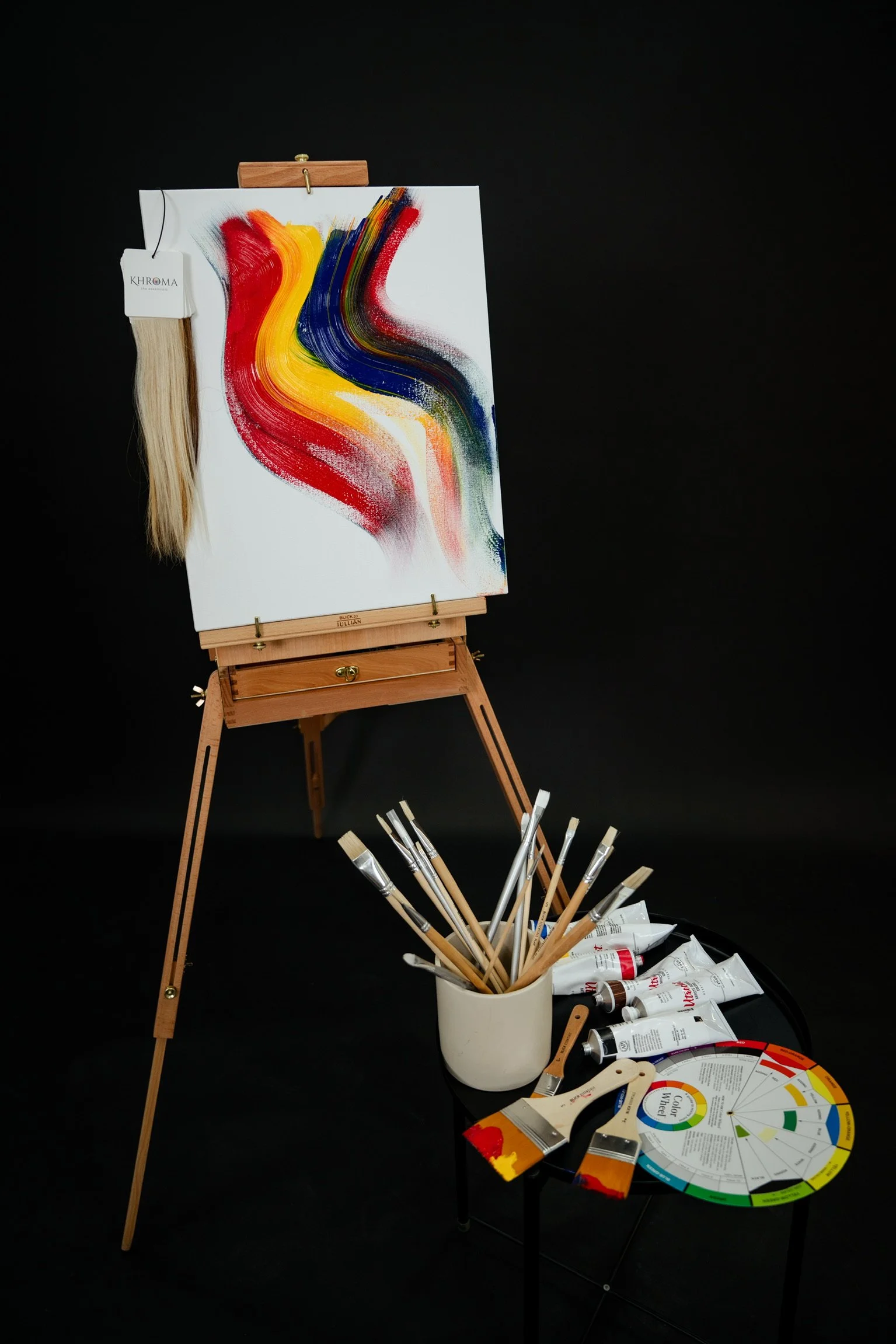 A painting easel with a colorful abstract painting, a palette, brushes, and paint tubes displayed against a black background.