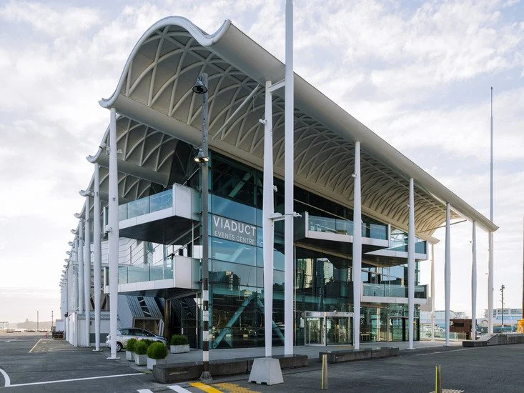 Modern glass building with white structural supports and a curved roof, labeled as Viaduct Events Centre, located in an outdoor area with parking spaces.