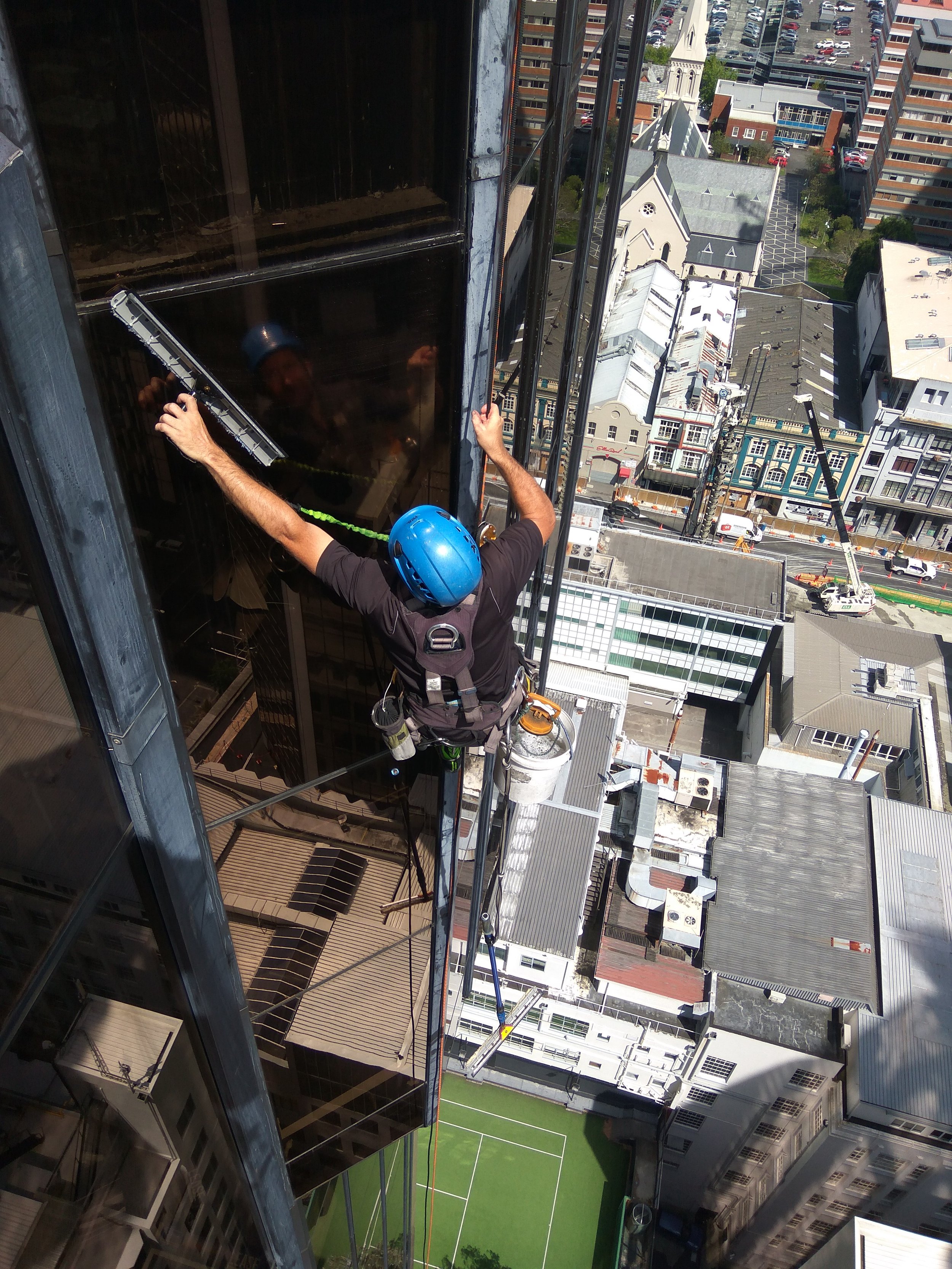 A window cleaner wearing a blue safety helmet and safety harness is cleaning the exterior glass of a high-rise building with a squeegee, with a cityscape and tennis court below.